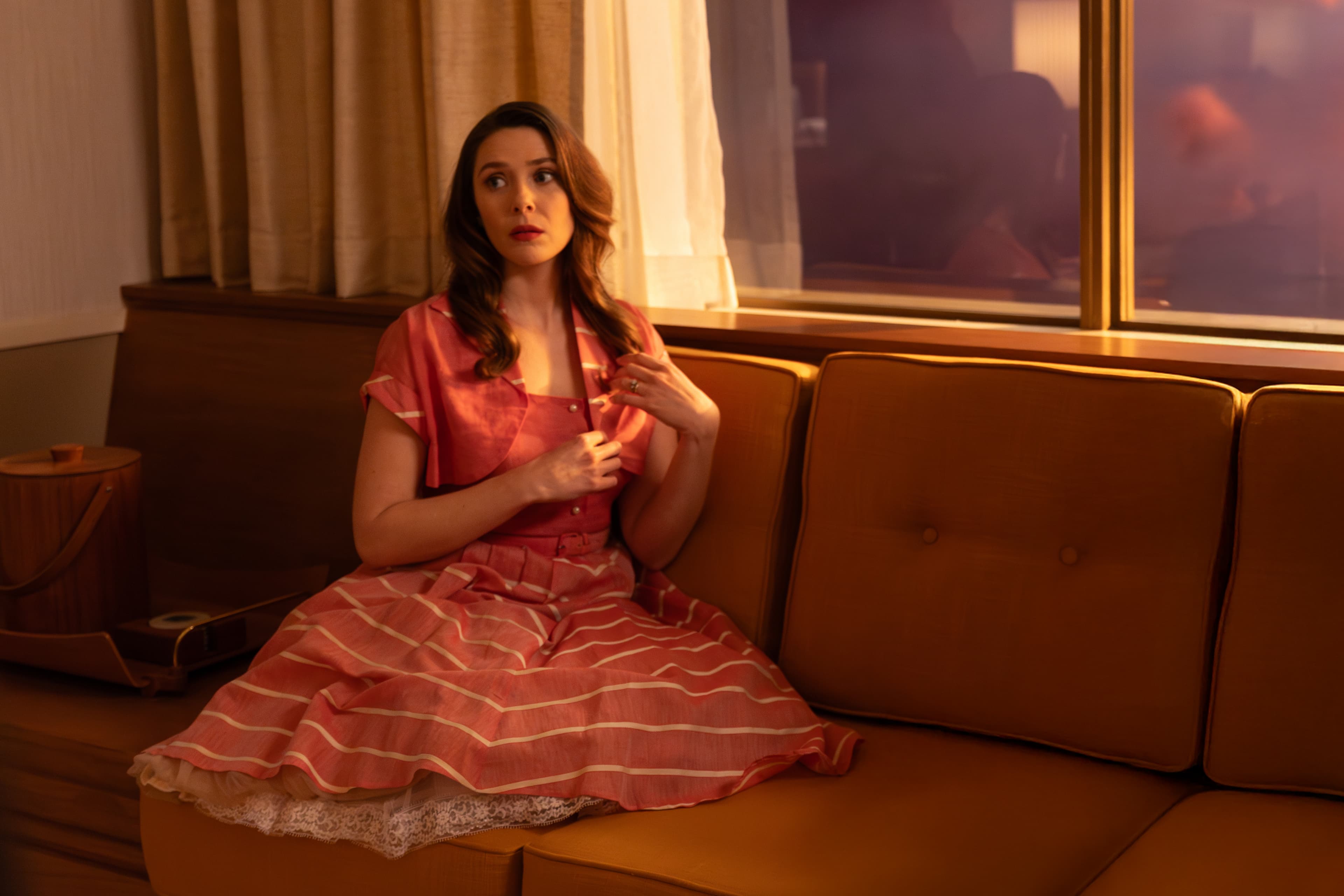 Actress Elizabeth Olsen sits on a couch in a '50s-style dress. 