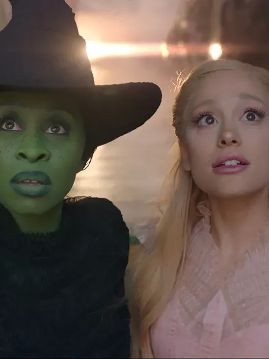 Cynthia Erivo as Elphaba, with green skin and a black witch-like ensemble, and Ariana Grande, as Glinda, dressed in a pink frilly dress, in a still from "Wicked." 