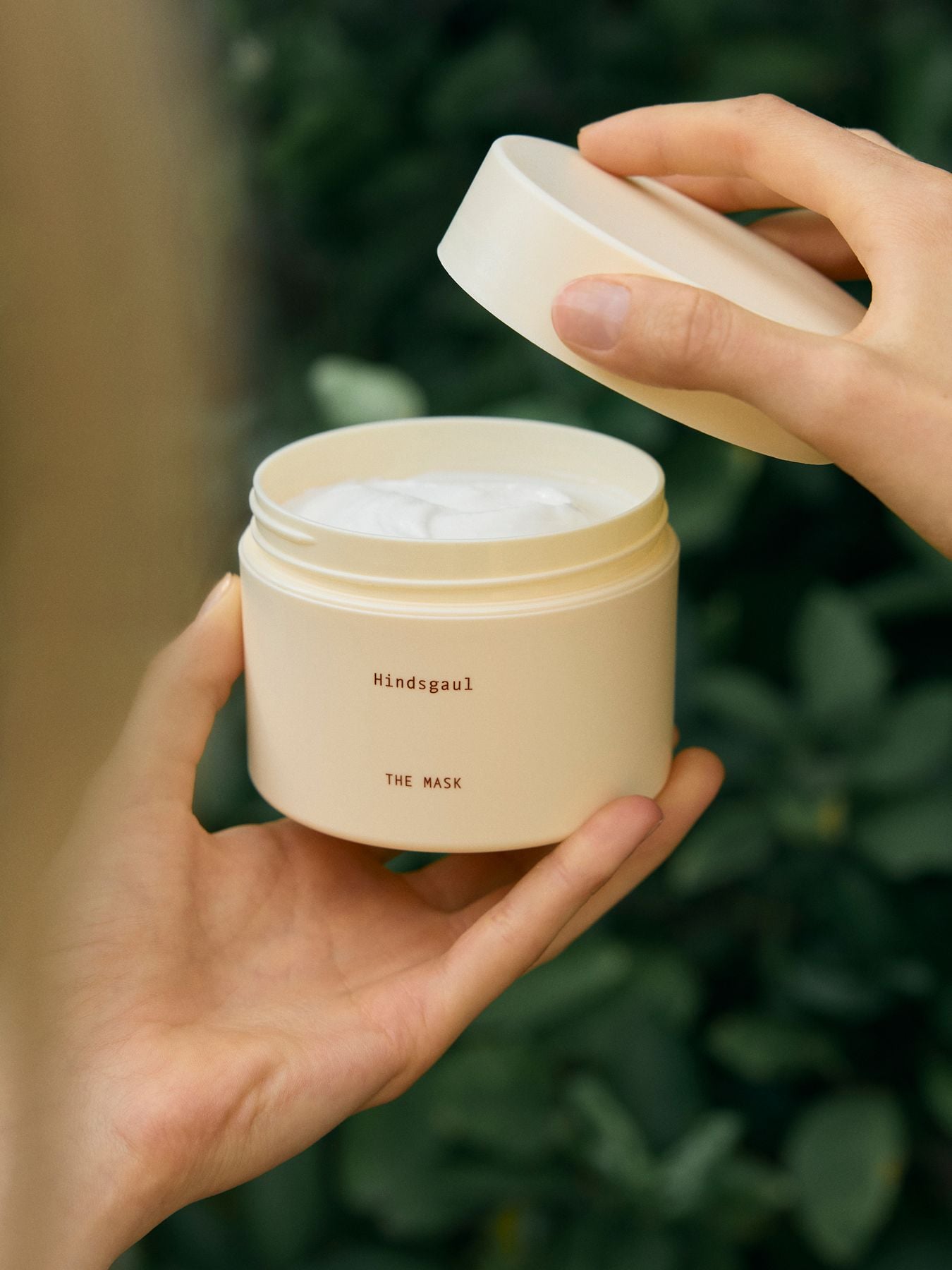 Hands opening a jar of The Hindsgaul Hair mask in front of a bush 