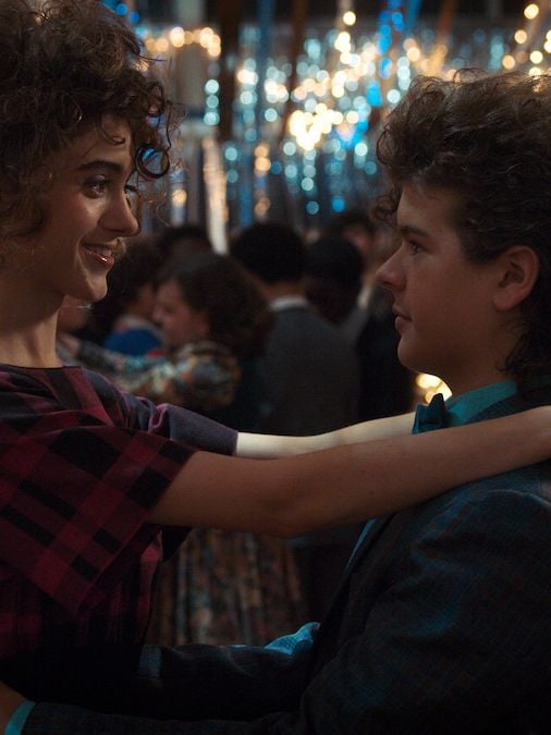 Actors Natalia Dyer and Gaten Matarazzo slow dance at a school dance, dressed in full 80s attire in a still from Stranger Things. 