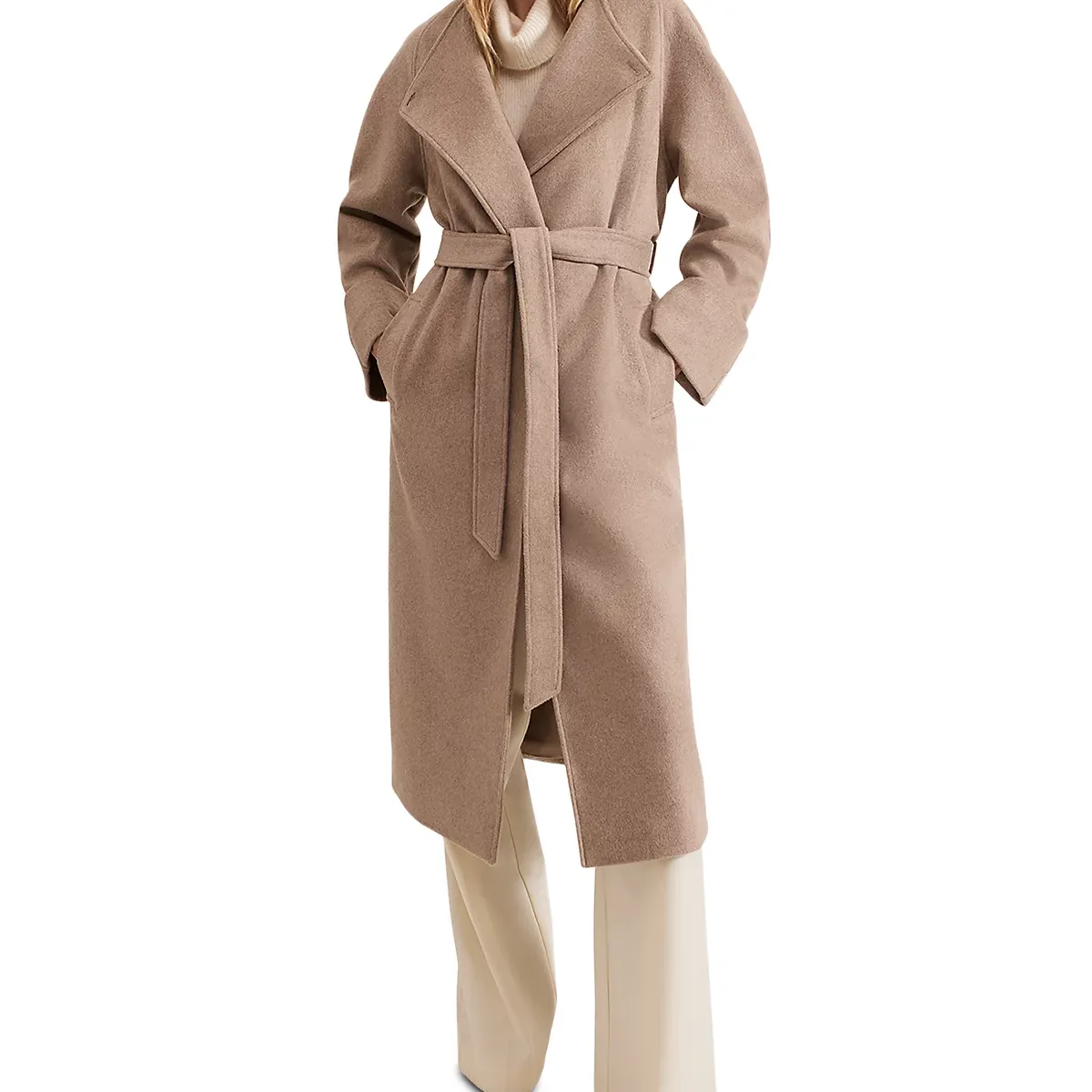 A model wearing a long beige coat with a belt and white trousers, standing against a white background 