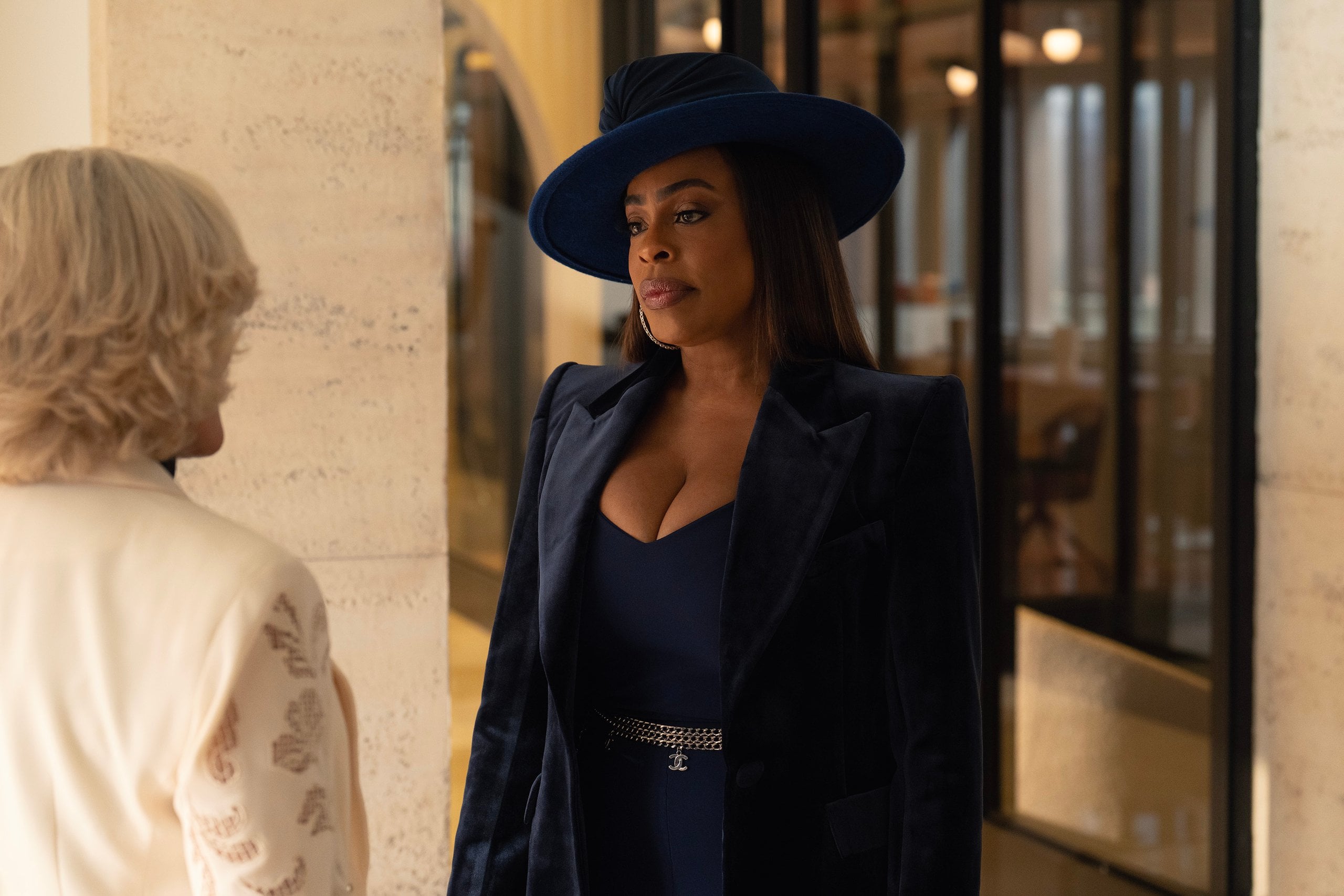 Niecy Nash stands in a lobby wearing an all-navy suit and hat in a still from All's Fair.