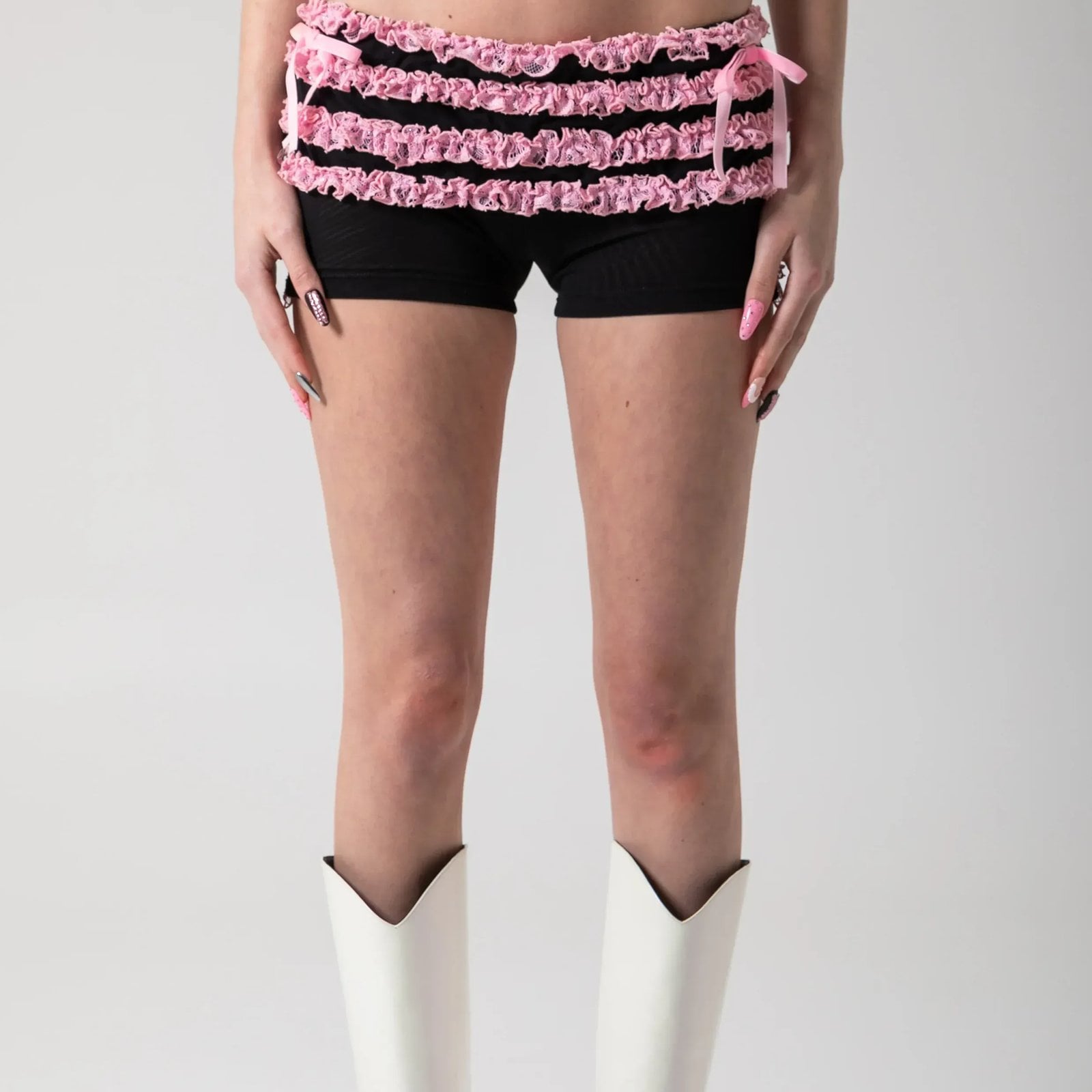 A model wearing black and pink ruffle shorts and white cowboy boots against a gray background