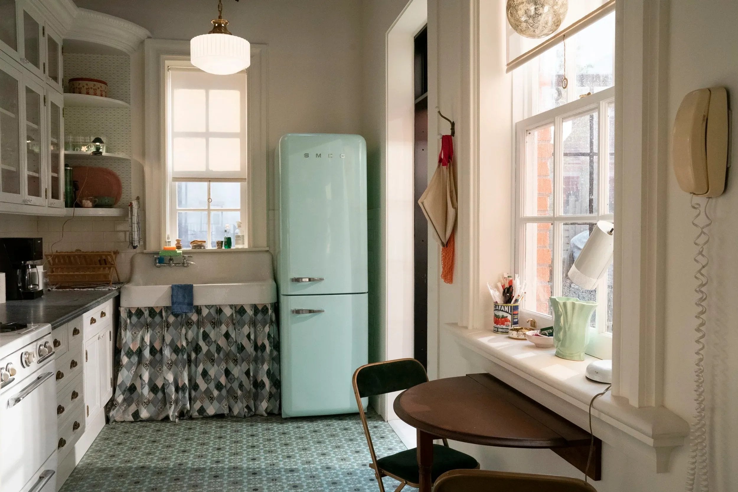 A photo of a kitchen, looking at the seafoam vintage-stle fridge, small halfmoon shaped wooden table, and window from the show "And Just Like That."