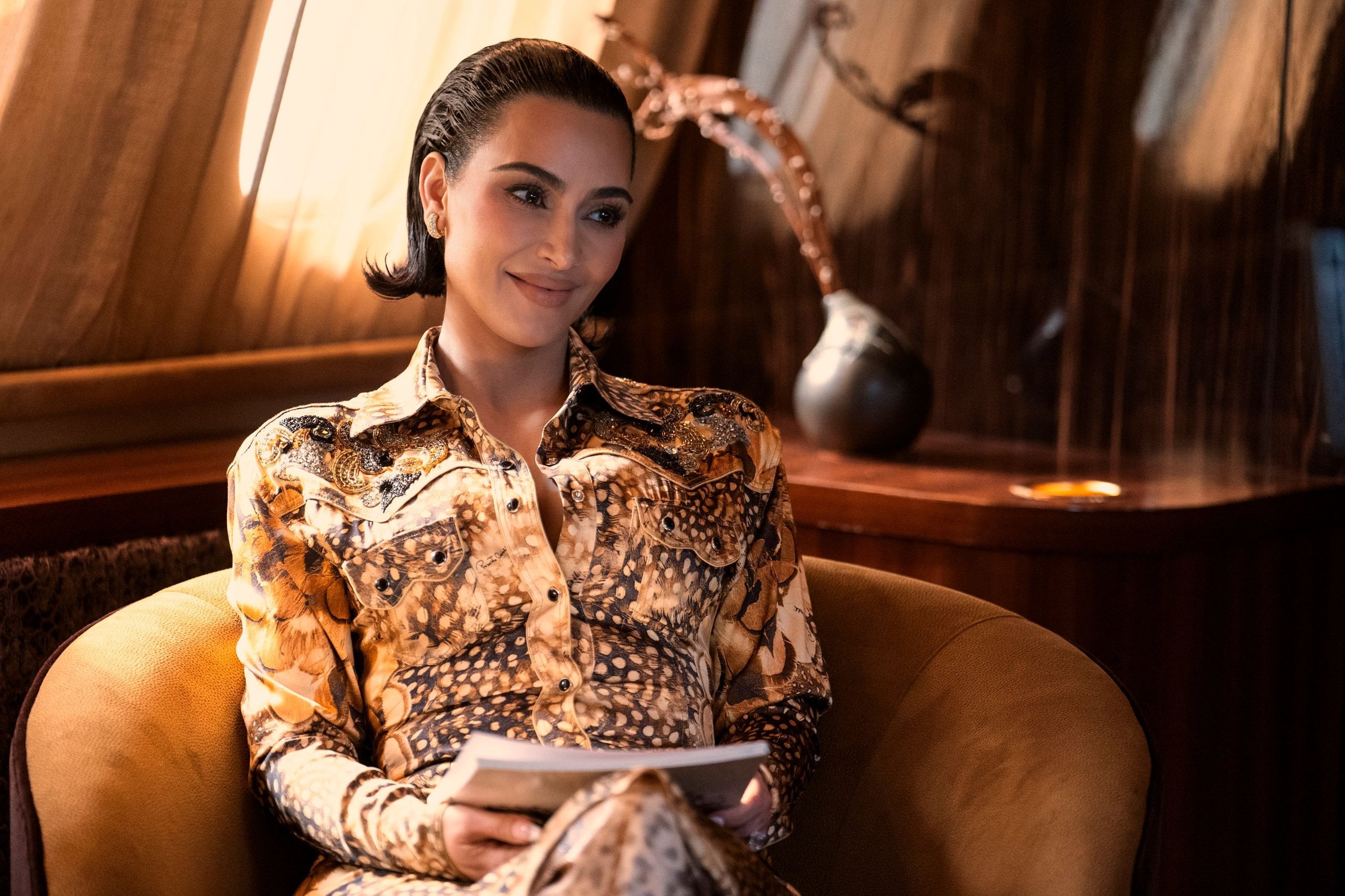 Kim Kardashian sits in a suede chair holding some papers in a still from Hulu's All's Fair.
