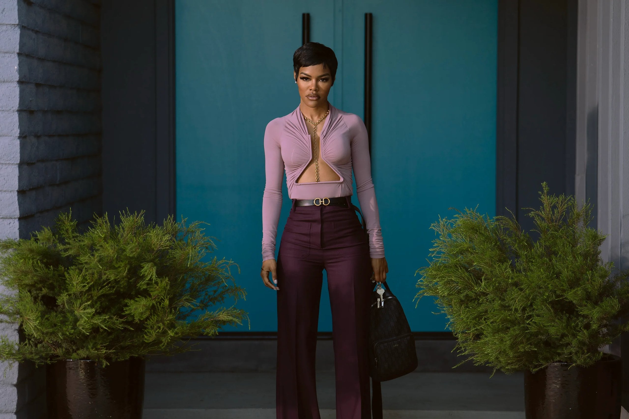 Actress Teyana Taylor stands in front a green walk in a pale lilac top and burgundy trousers in a still from Hulu's All's Fair.