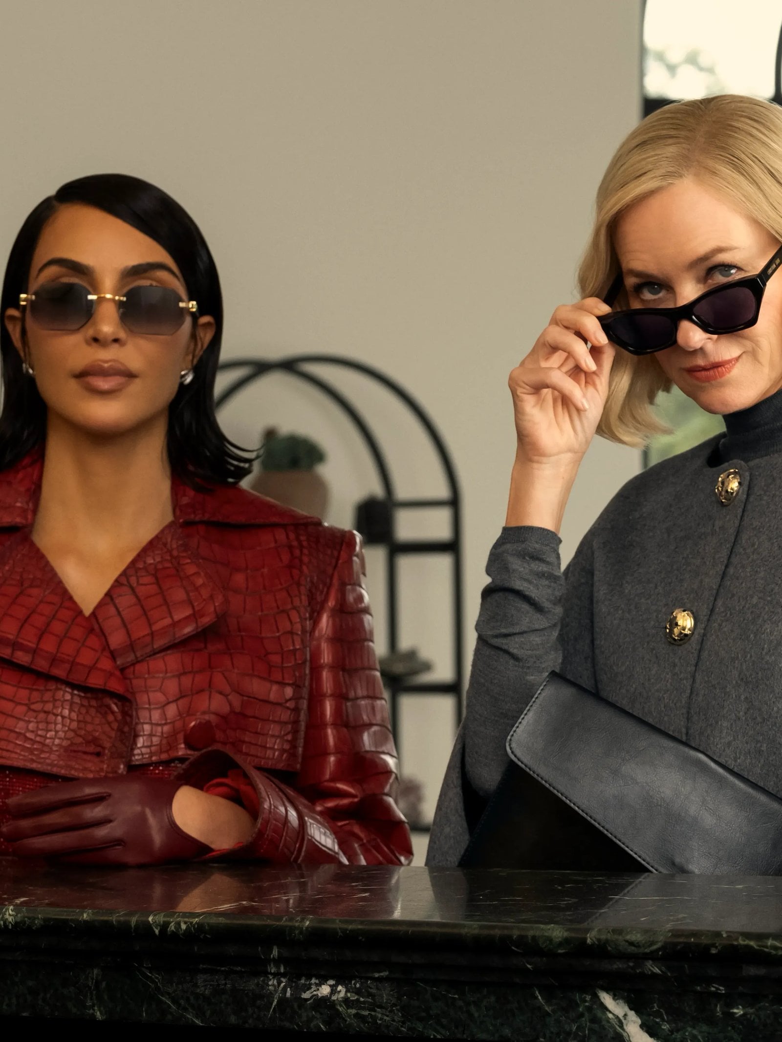 Kim Kardashian and Naomi Watts stand in front of a desk in sunglasses and suits in a still from Hulu's All's Fair.