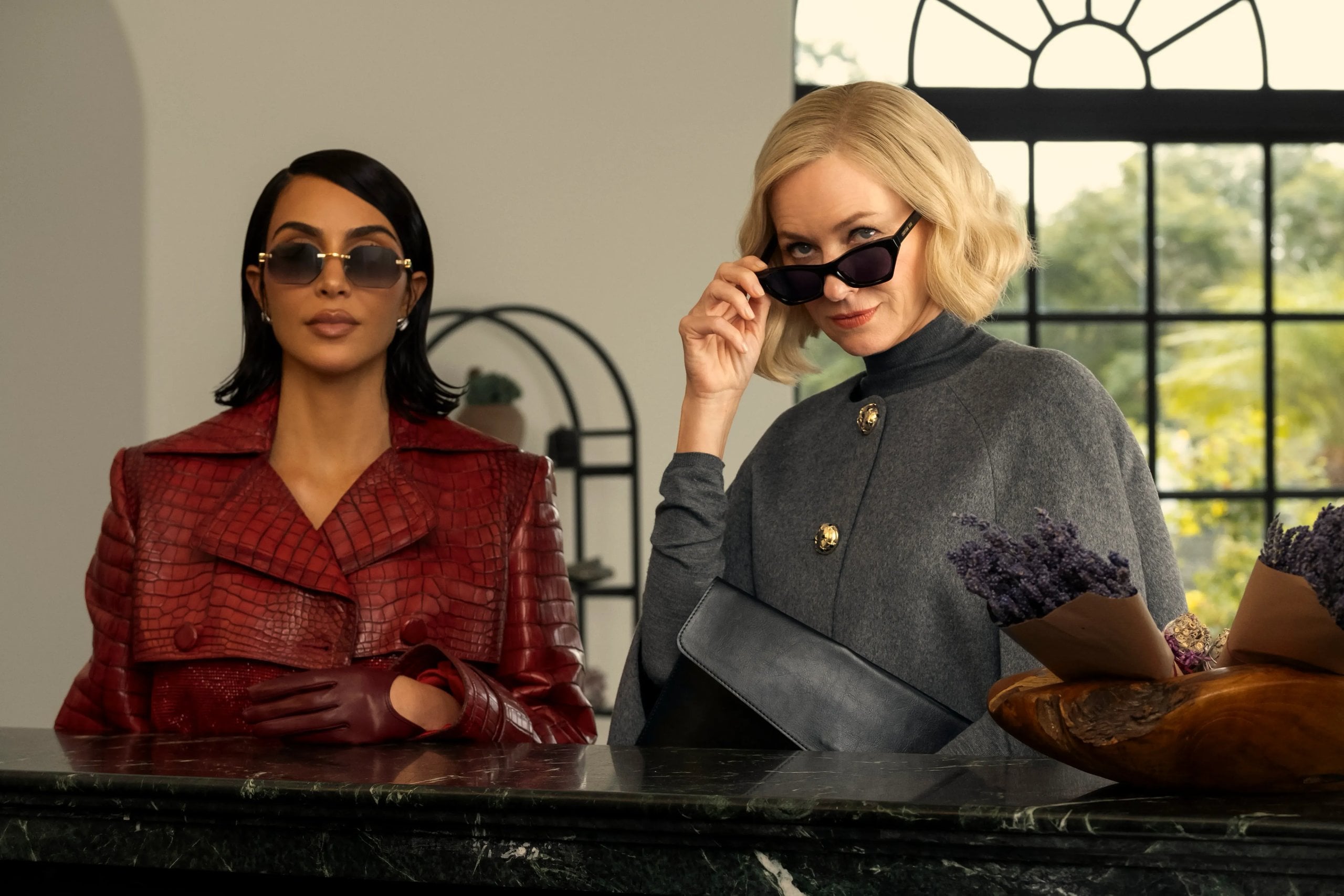 Kim Kardashian and Naomi Watts stand in front of a desk in sunglasses and suits in a still from Hulu's All's Fair.