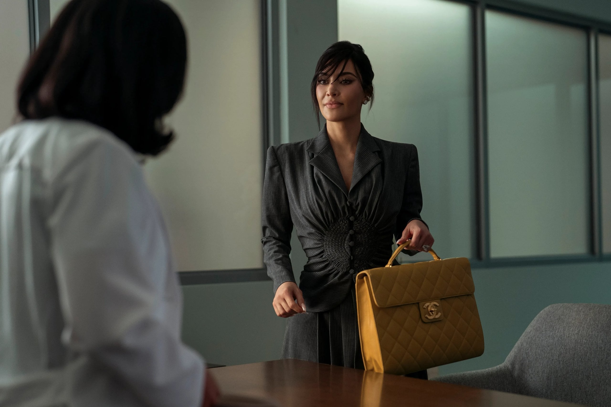 Kim Kardashian stands in an office in a gray suit and holding a yellow Chanel briefcase in a still from Hulu's All's Fair.