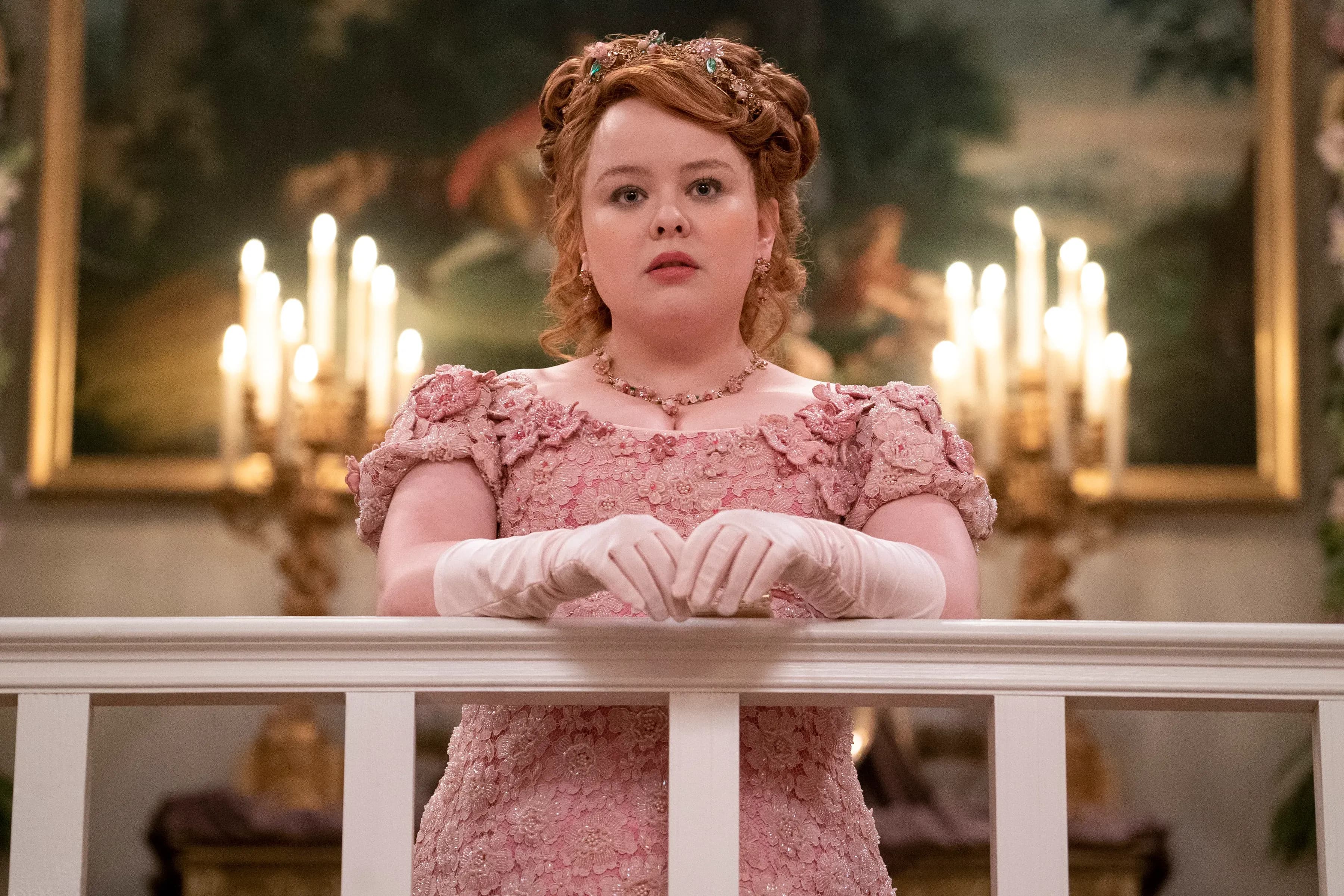 Nicola Coughlan wears a sparkling pink dress and pale-pink gloves, standing in front of two candelabras in a scene from Bridgerton.