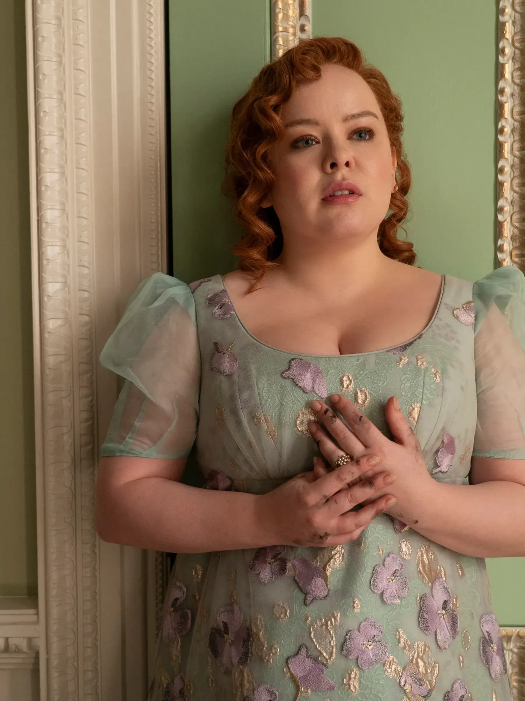 Actress Nicola Coughlan wears a seafoam-green dress and stands agains a green-and-gold wall in a scene from Bridgerton.