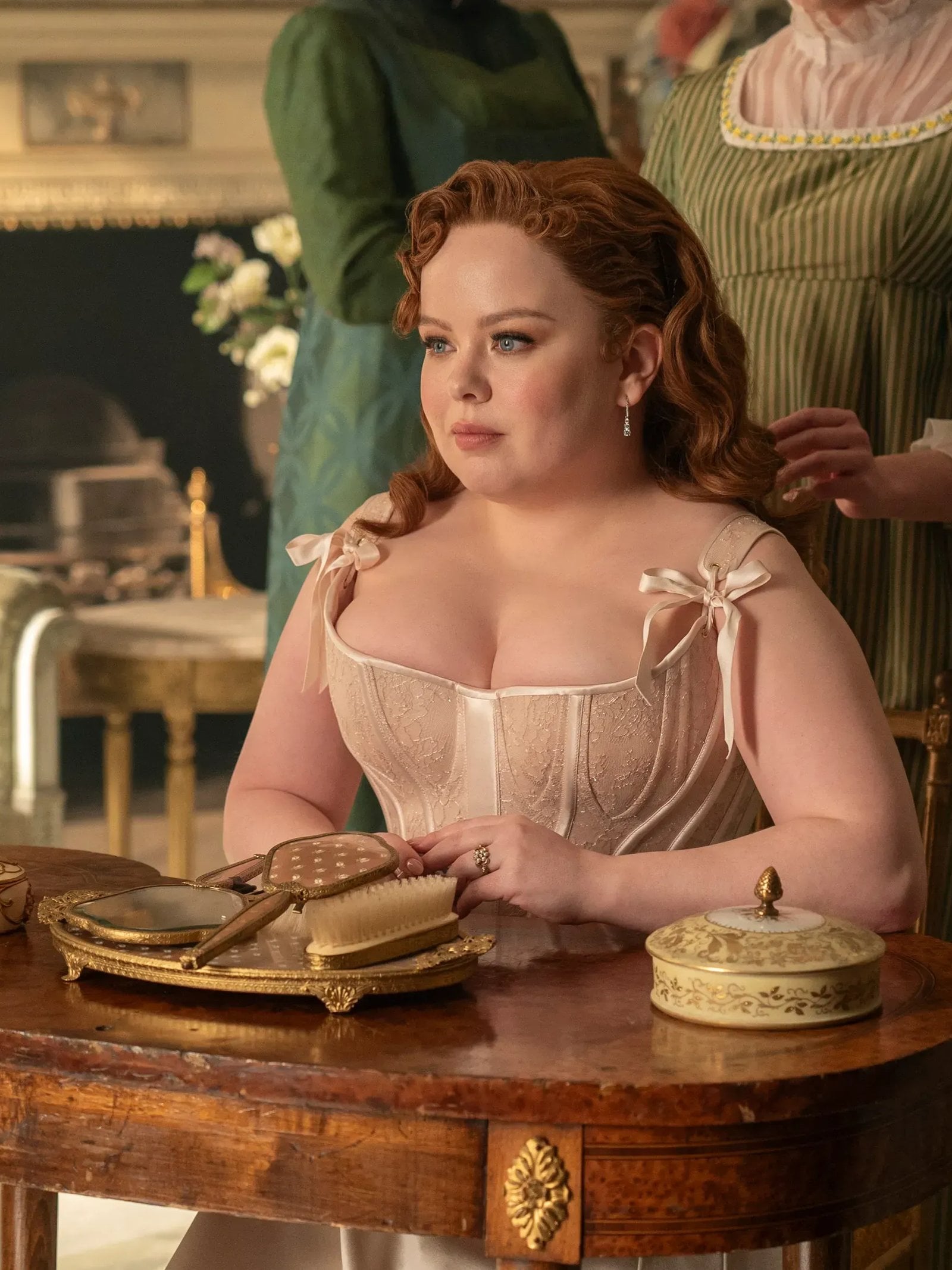 Nicola Coughlan sits at a table with her arms folded, wearing a beige corset dress.
