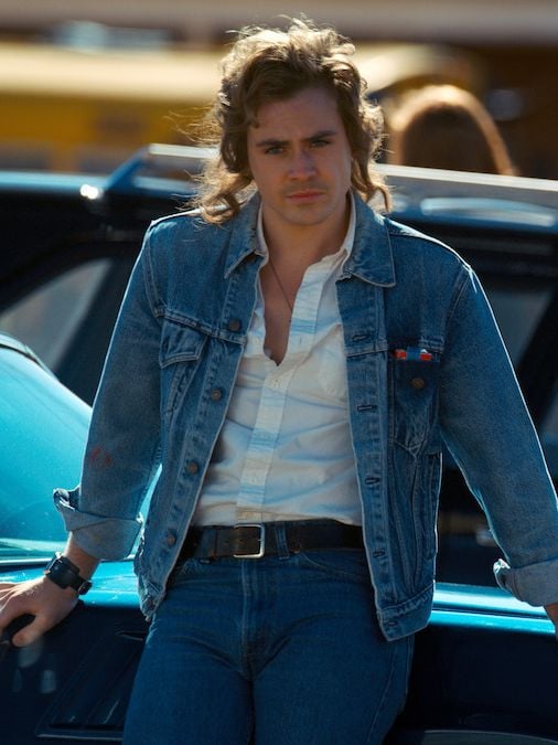 Actor Dacre Montgomery with a mullet, wearing a white t-shirt and jean jacket in a still from Stranger Things.