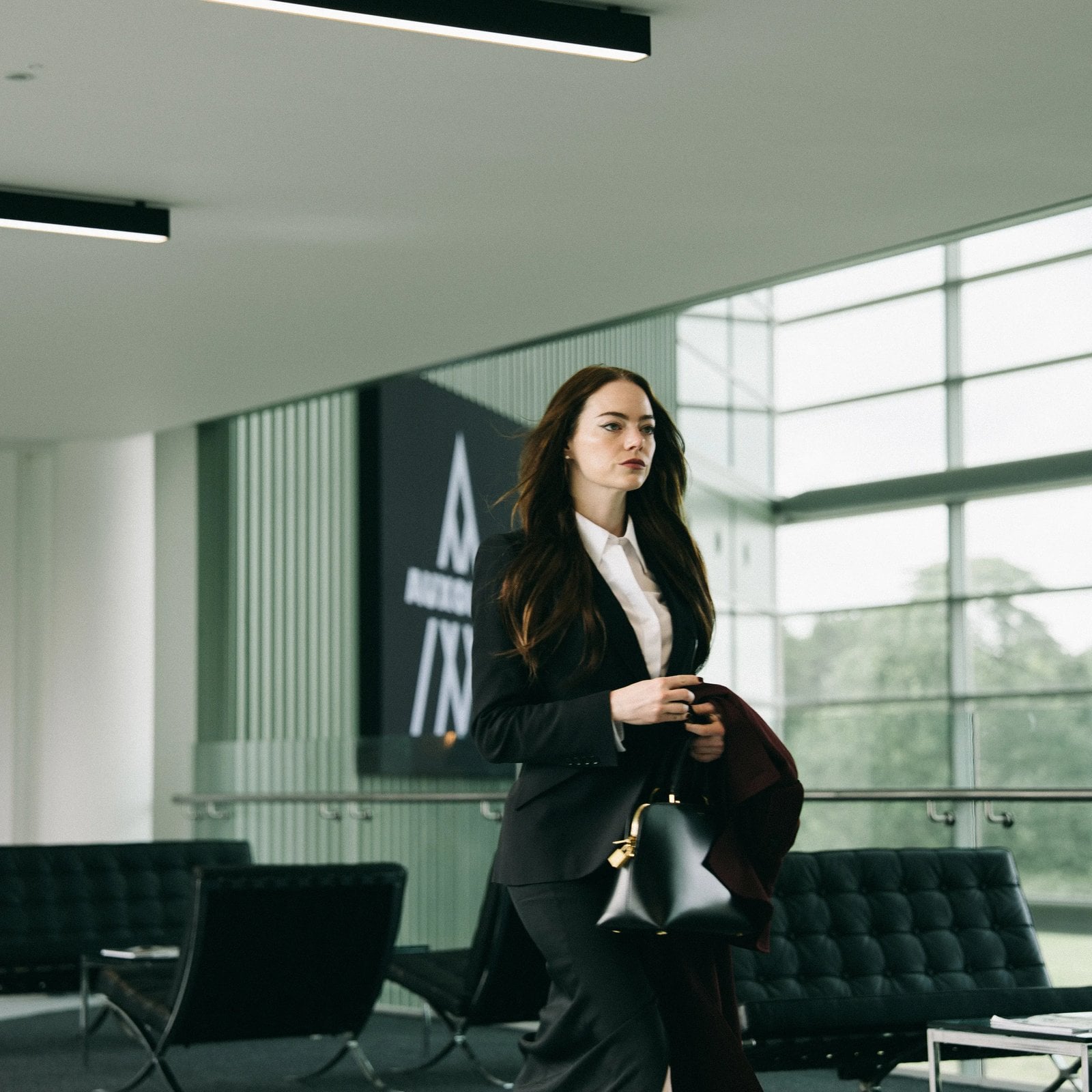 Actor Emma Stone stands in a modern office wearing a suit and carrying a black bag in a still from "Bugonia."