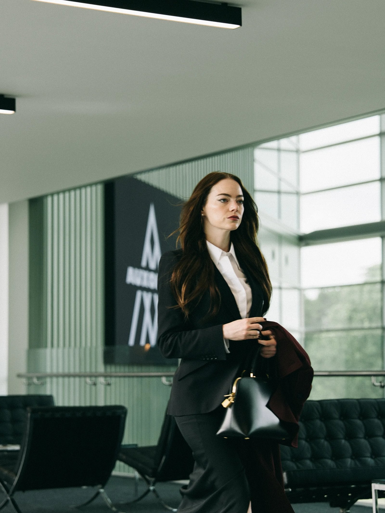 Actor Emma Stone stands in a modern office wearing a suit and carrying a black bag in a still from "Bugonia."