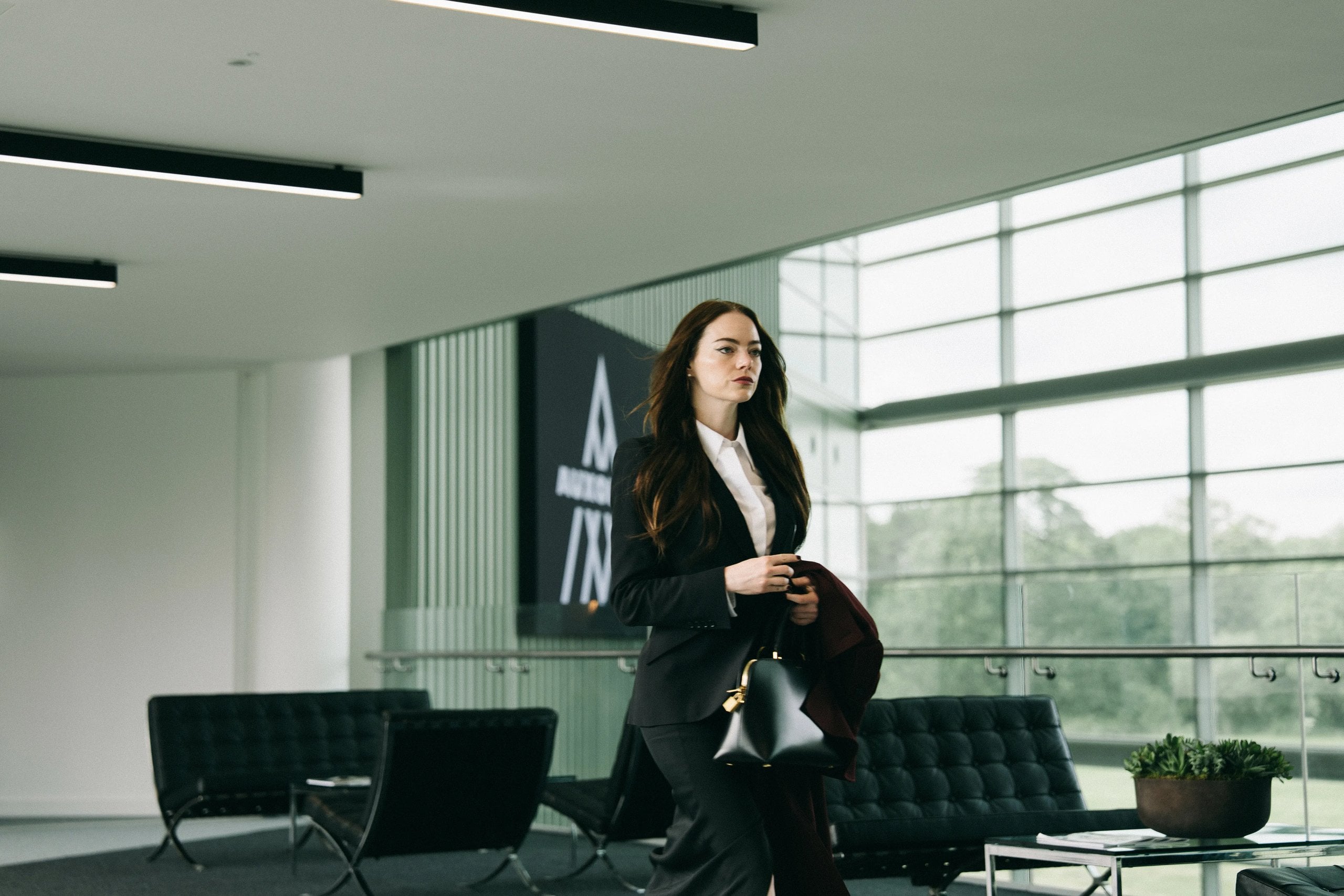 Actor Emma Stone stands in a modern office wearing a suit and carrying a black bag in a still from "Bugonia."