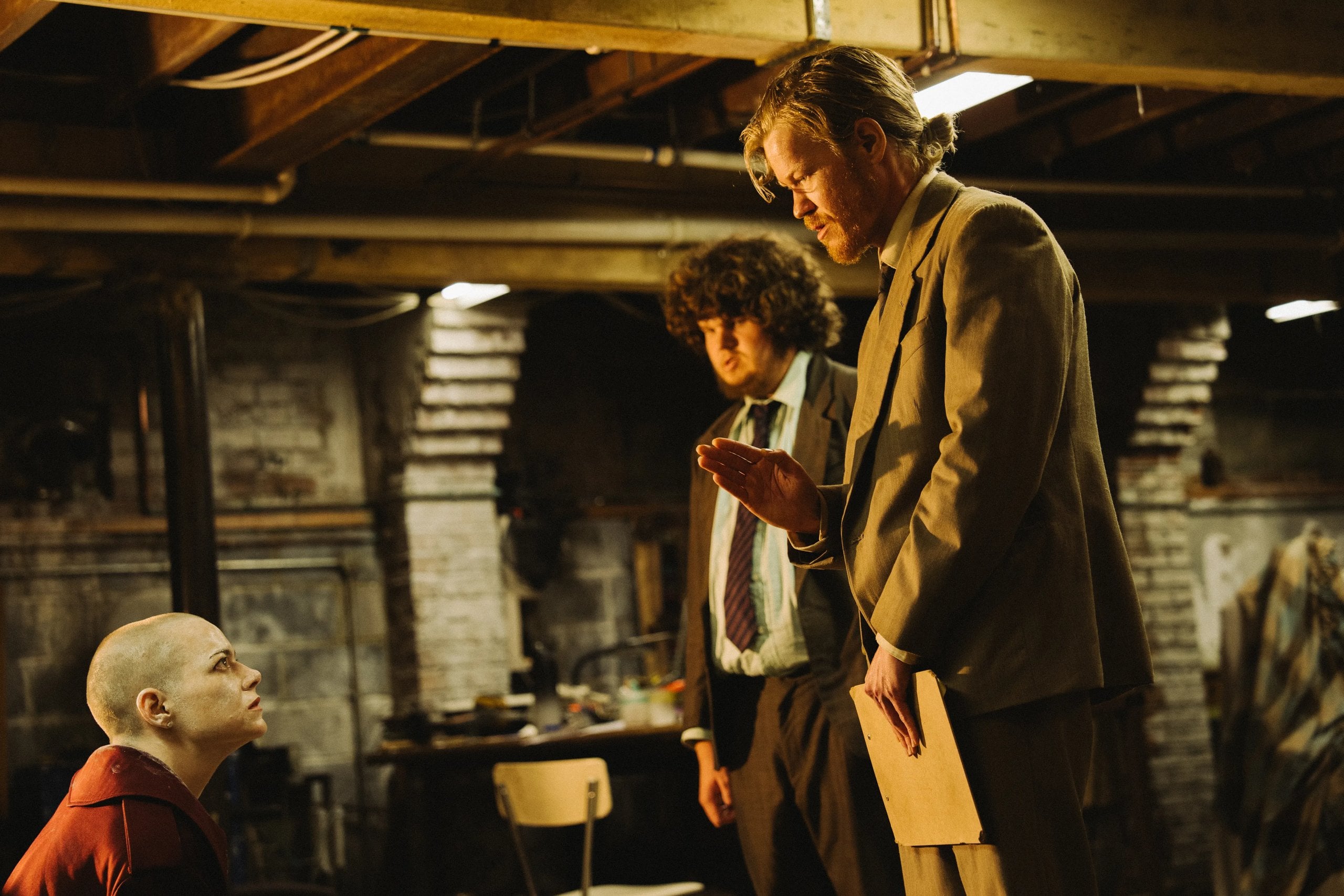 Actors Aidan Delbis and Jesse Plemons stand together talking to someone off screen in a still from "Bugonia."