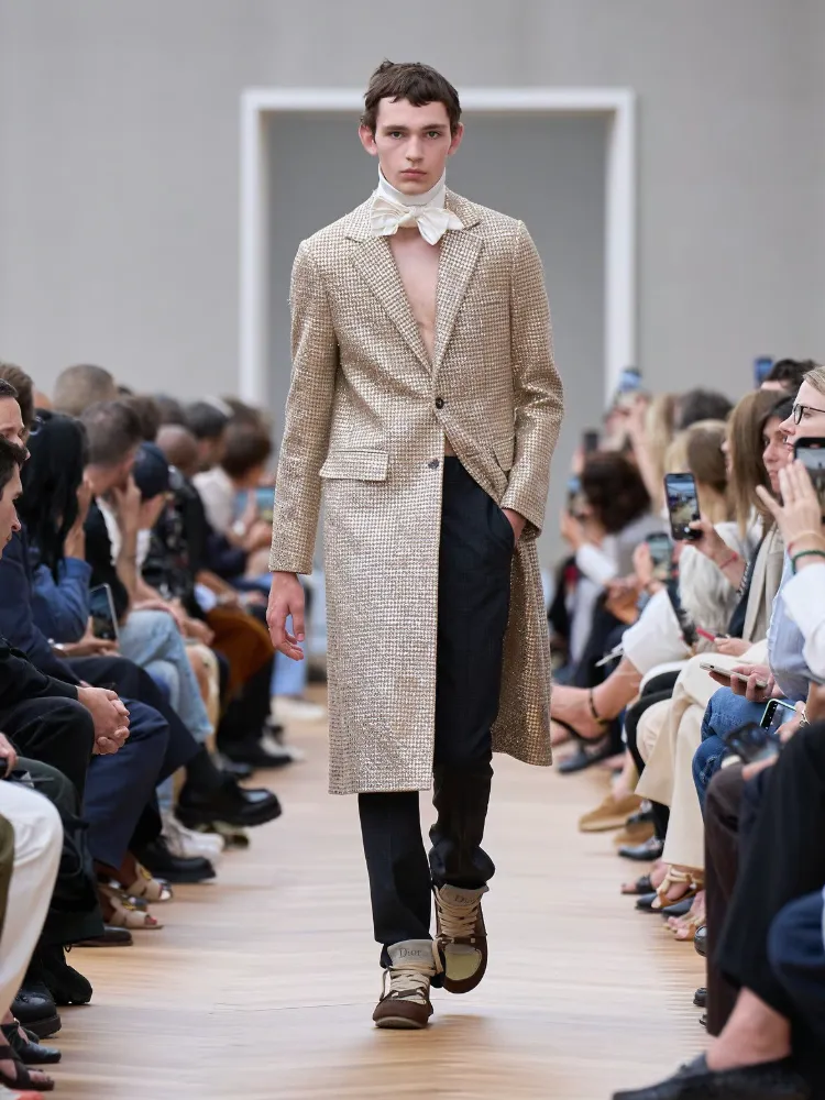 A male model walks down the Christian Dior Spring/Summer 2026 runway in a long gilded coat, white cravat, and black trousers.