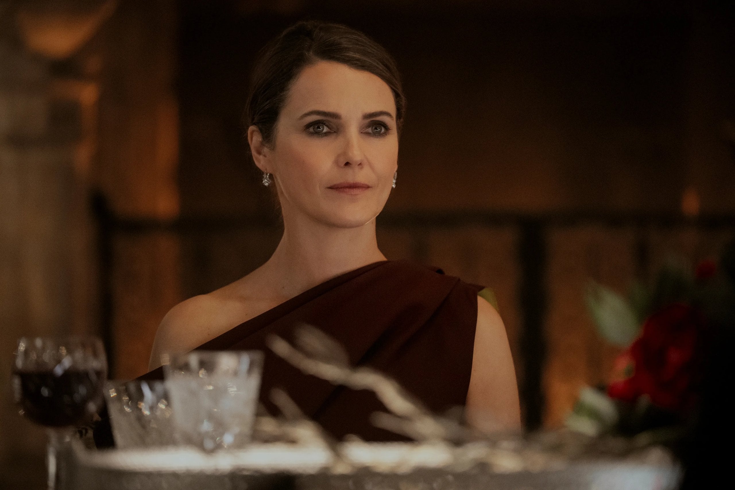 Actress Keri Russell sits at a dinner table in one shoulder dress in a still from The Diplomat.