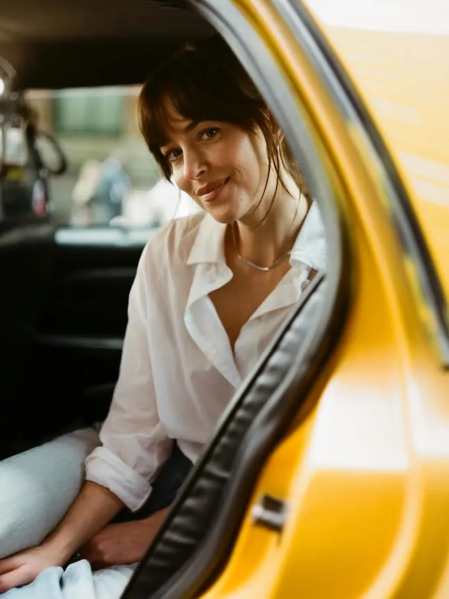 Dakota Johnson sits inside a New York City yellow taxi cab wearing a white shirt and jeans