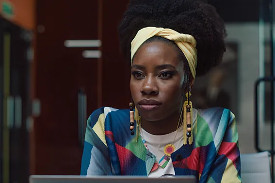 Actor Dani Moseley sits at a desk wearing a graphic sweater and a yellow headband looking serious in a still from "Dreaming Whilst Black."