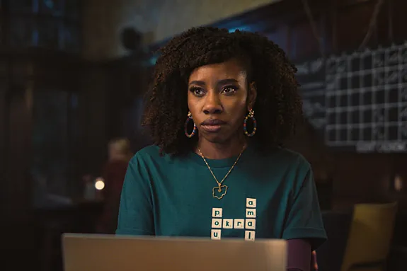 Actor Dani Moseley sits at a desk wearing a green sweater with a crossword design on it, in a still from "Dreaming Whilst Black."