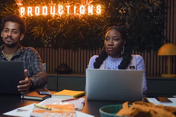 Two actors sit at a table with their laptops, in front light-up sign that says "productions" in a scene from Dreaming Whilst Black.