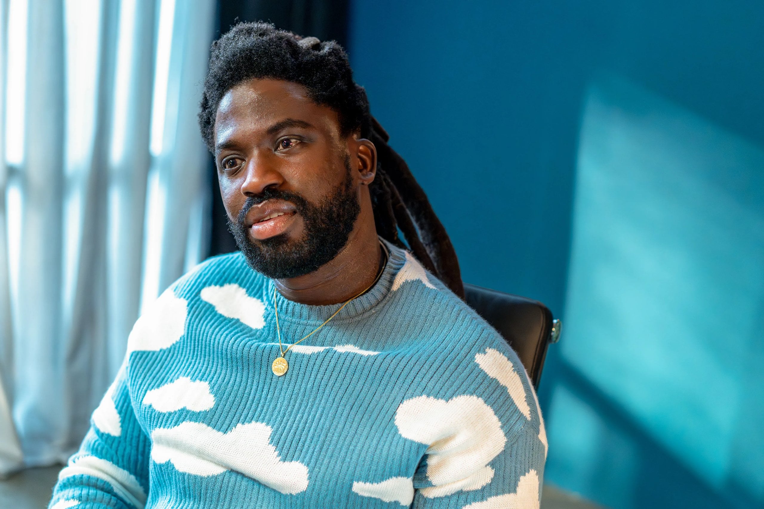 Actor Adjani Salmon sits in front of a blue wall, wearing a blue sweater with white clouds, his head tilted to the left.