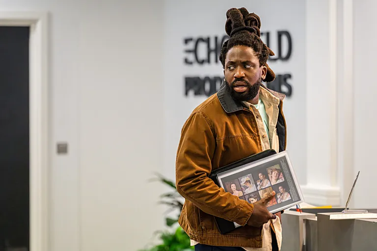 Actor Adjani Salmon wear a tan cordory jacket and holding a binder, in a still from "Dreaming Whilst Black."