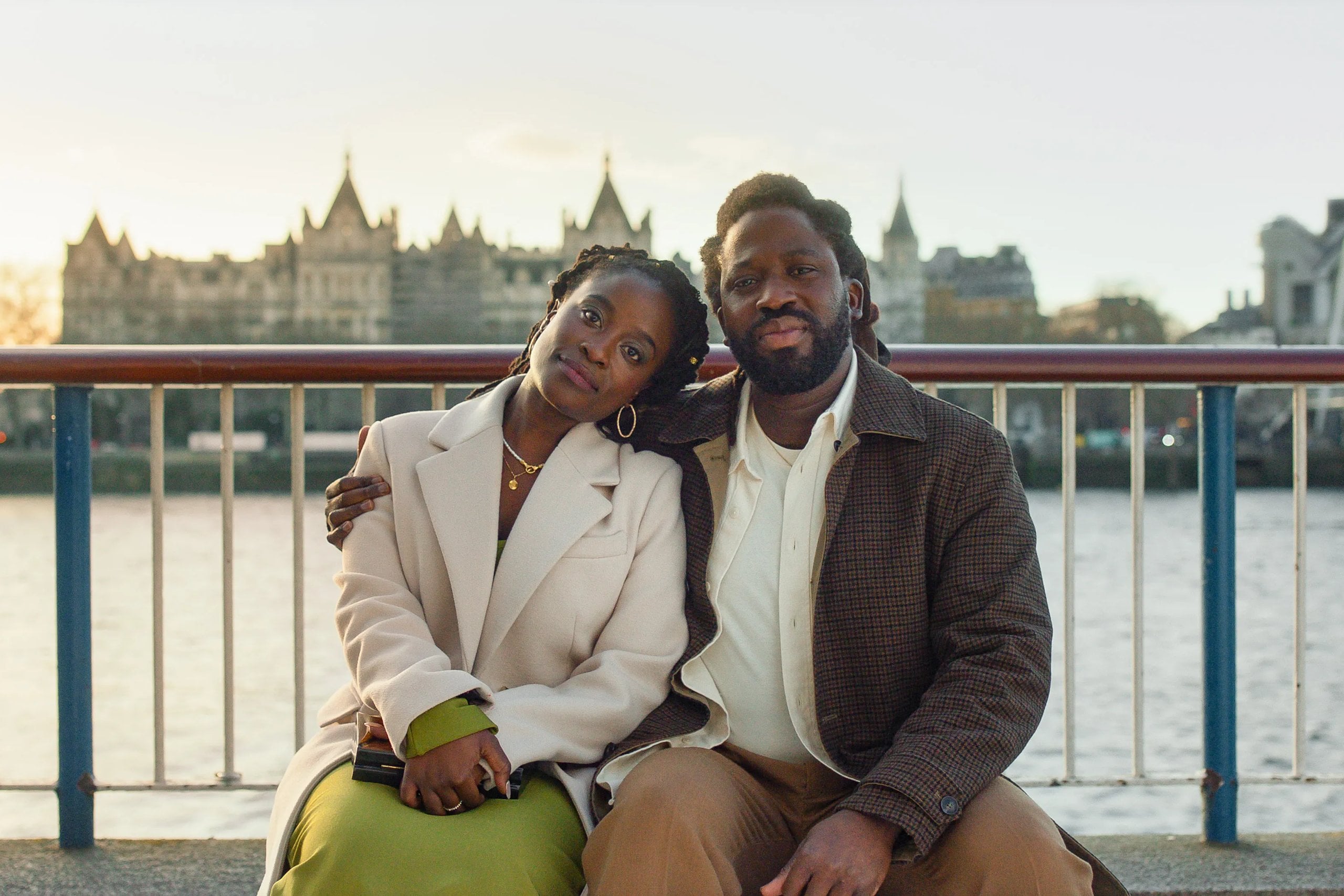Actors Adjani Salmon and Babirye Bukilwa sit on a bench in front of the Thames River in a still from "Dreaming Whilst Black."