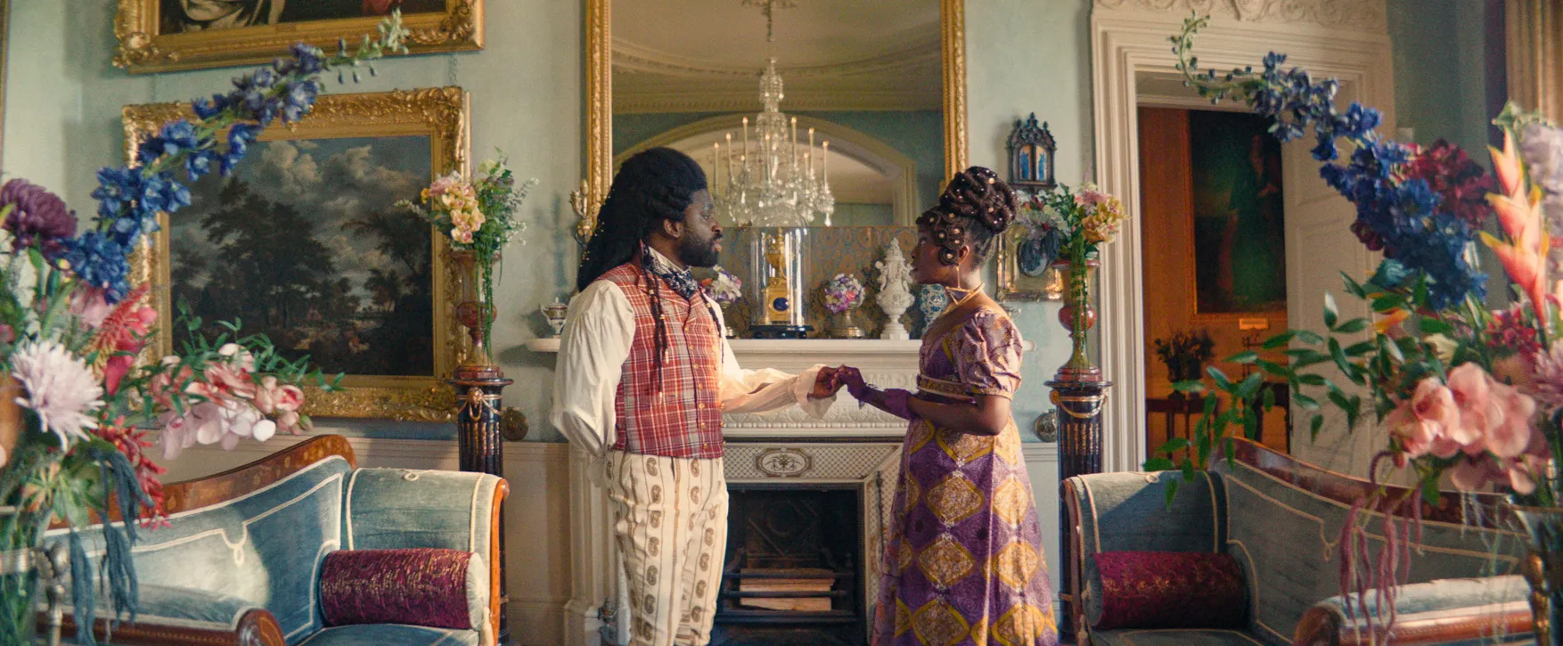 Actors Adjani Salmon and Babirye Bukilwa stand in a grand room in front of a mirror, holding hands and dressed in Regency-era clothing, in a still from "Dreaming Whilst Black."