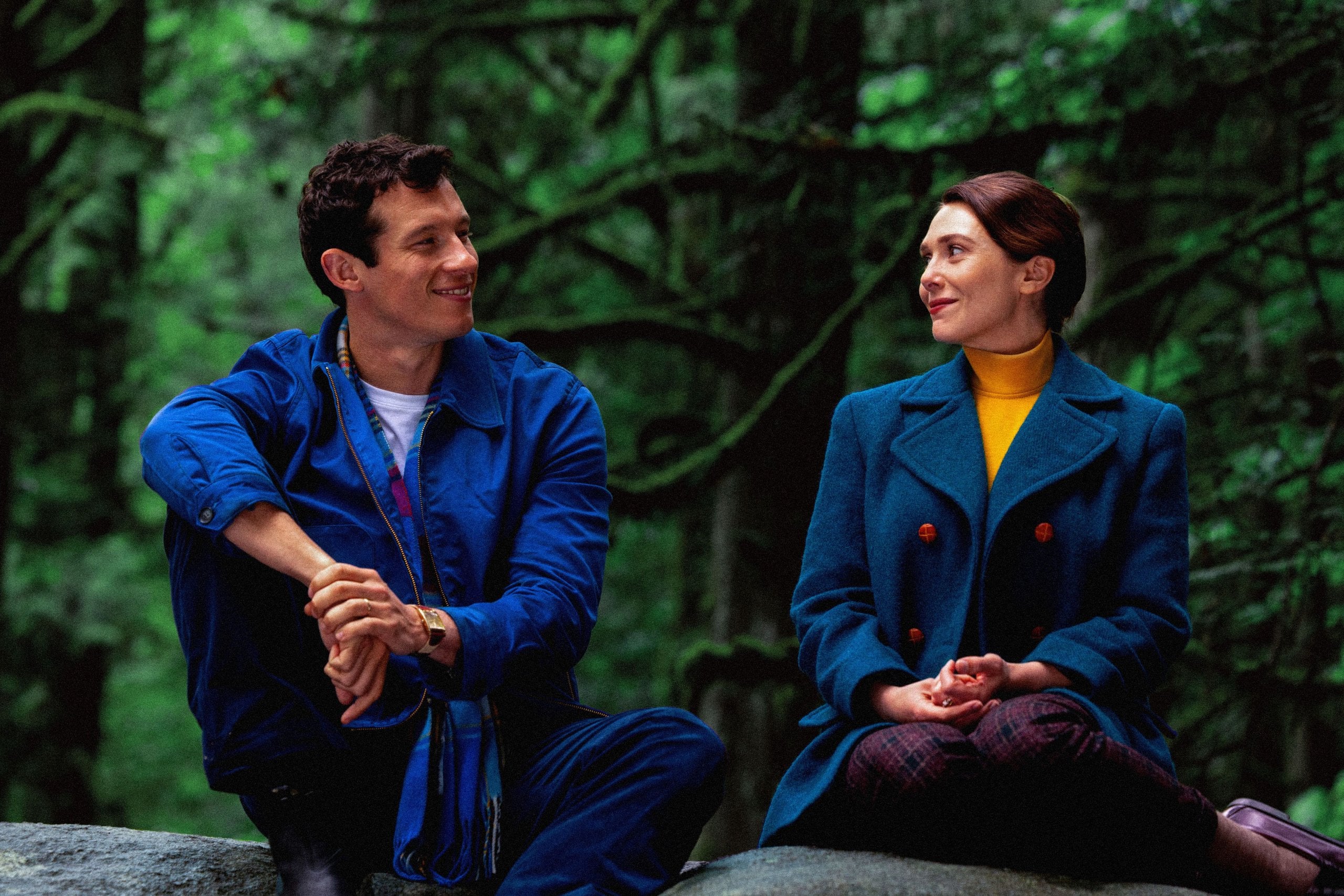 Actors Callum Turner and Elizabeth Olsen sit in the woods smiling at each other in a still from Eternity.