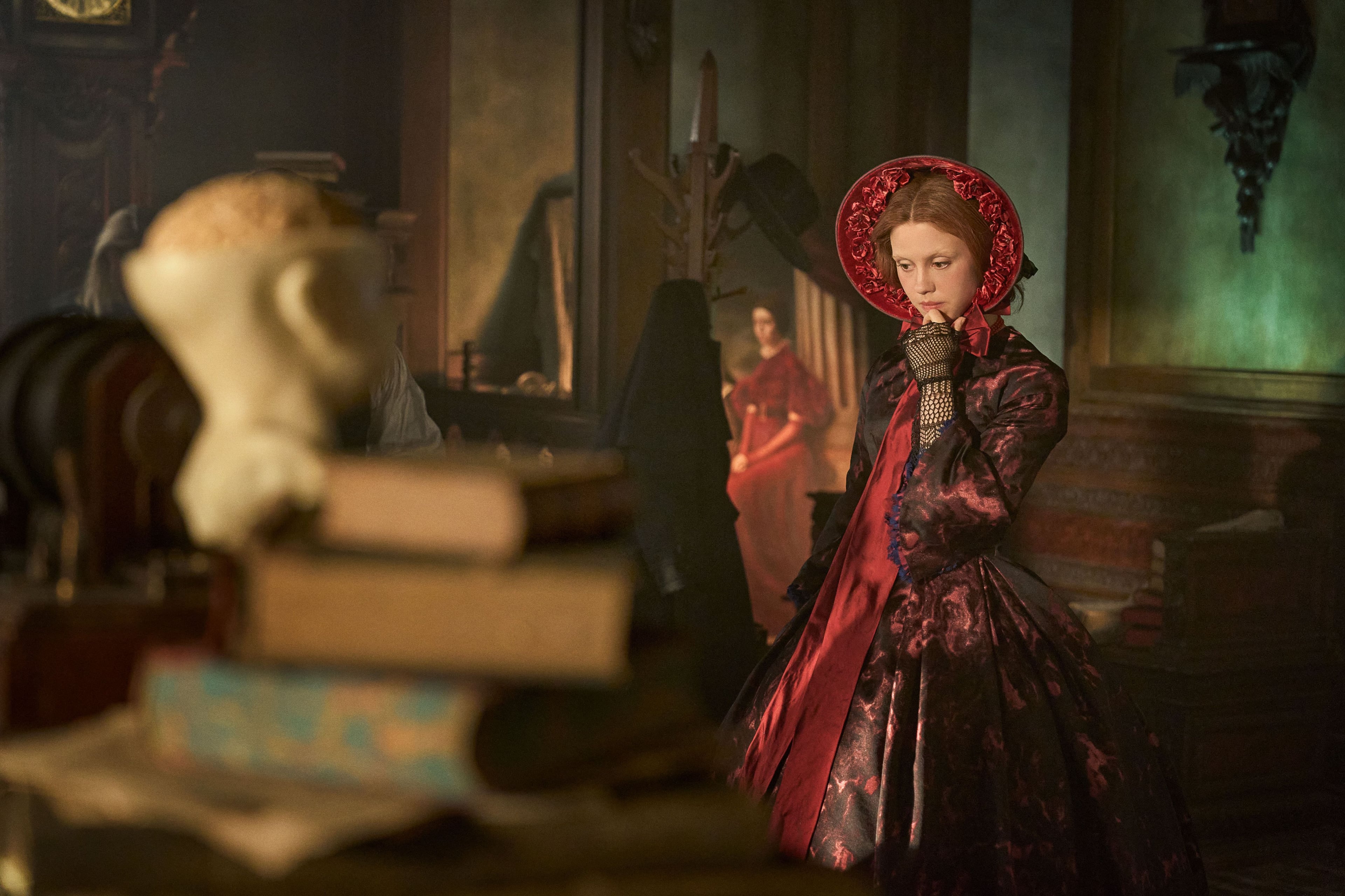 Actress Mia Goth stands in a dusty, dark room wearing a red dress and bonnet in a still from Frankenstein.