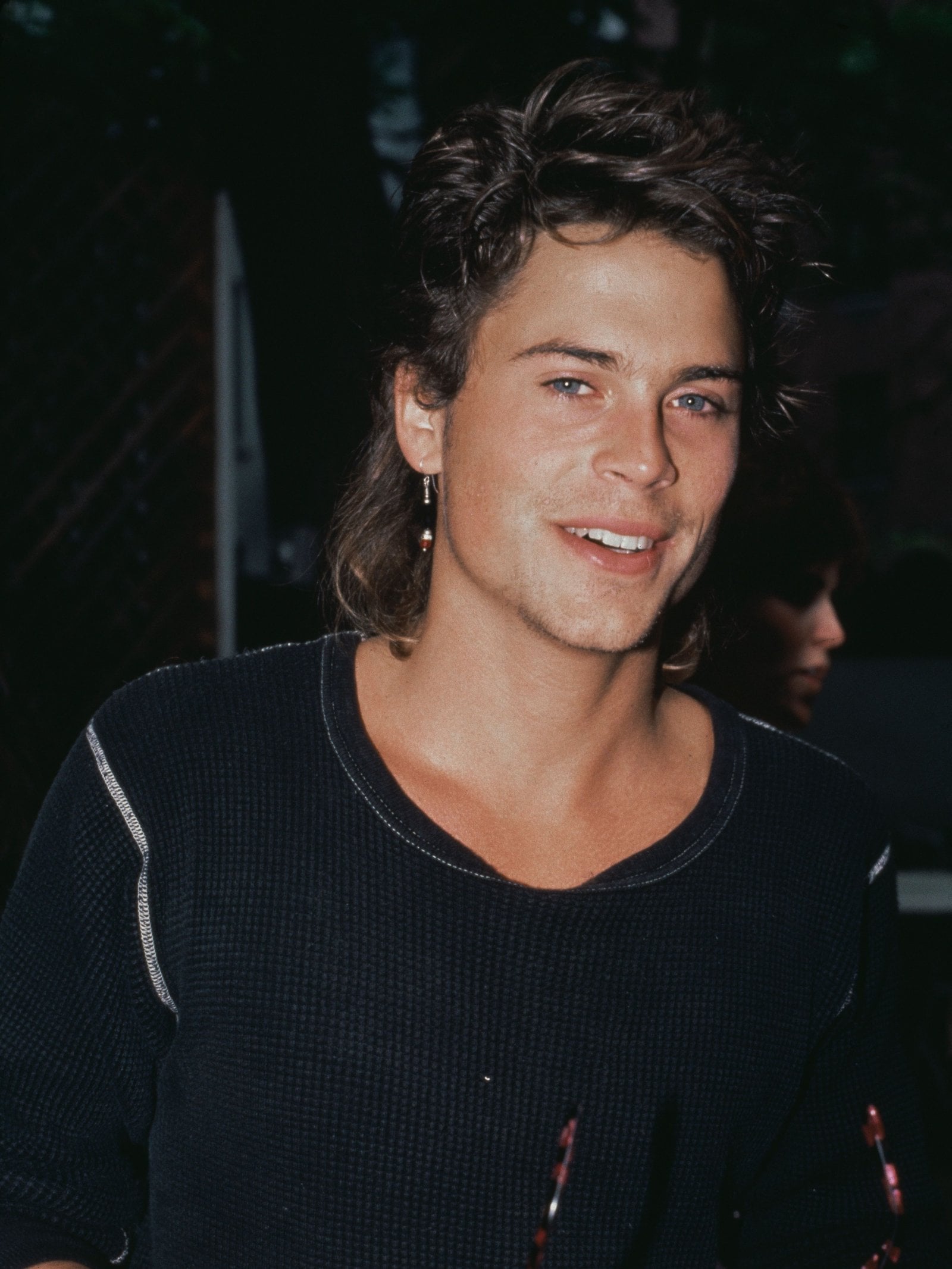 American actor Rob Lowe wearing a dark crew neck top with white trim, and holding a pair of sunglasses with tortoiseshell frames, circa 1985.