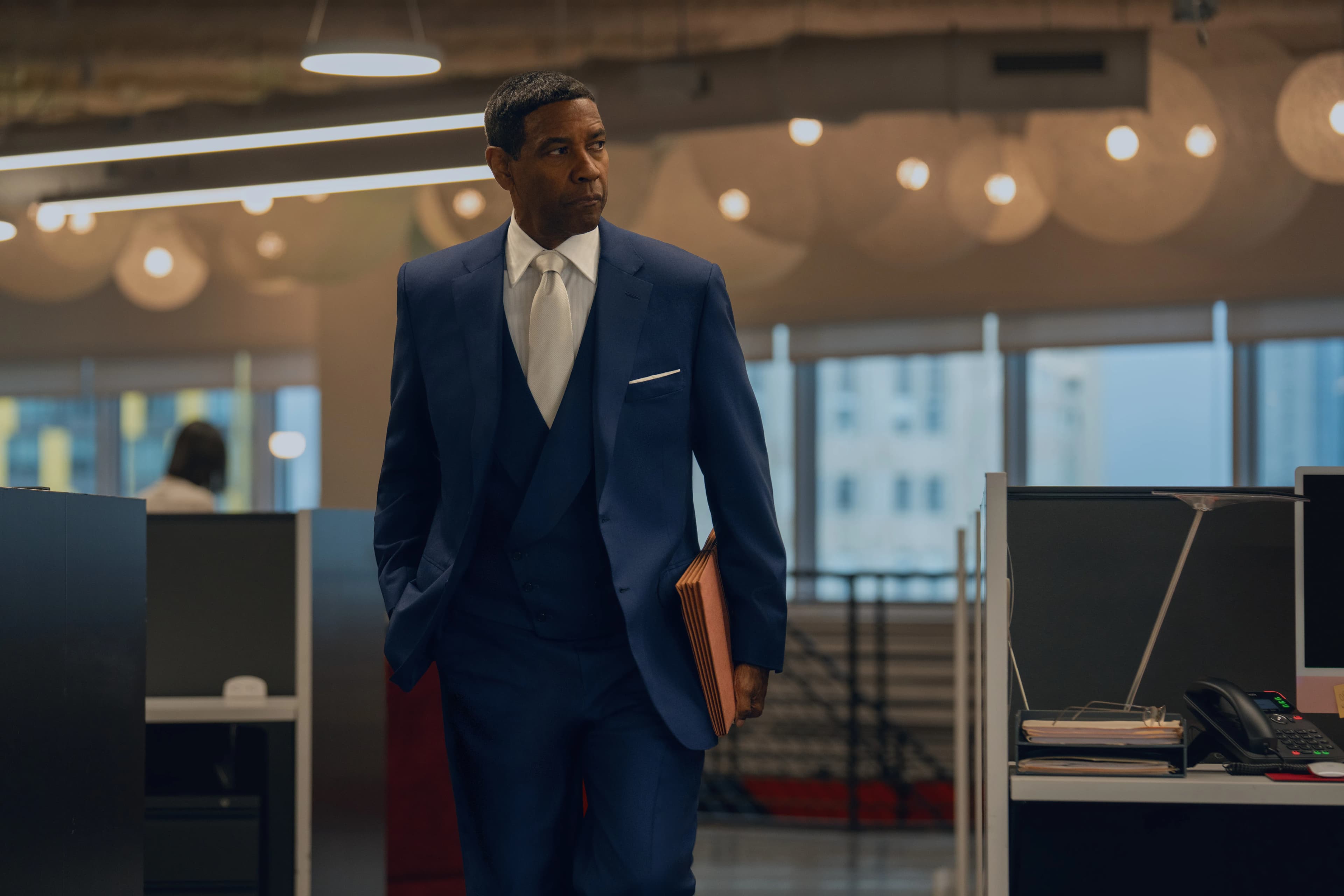 Actor Denzel Washington stands in an office wearing a navy blue suit and carrying a tan briefcase.
