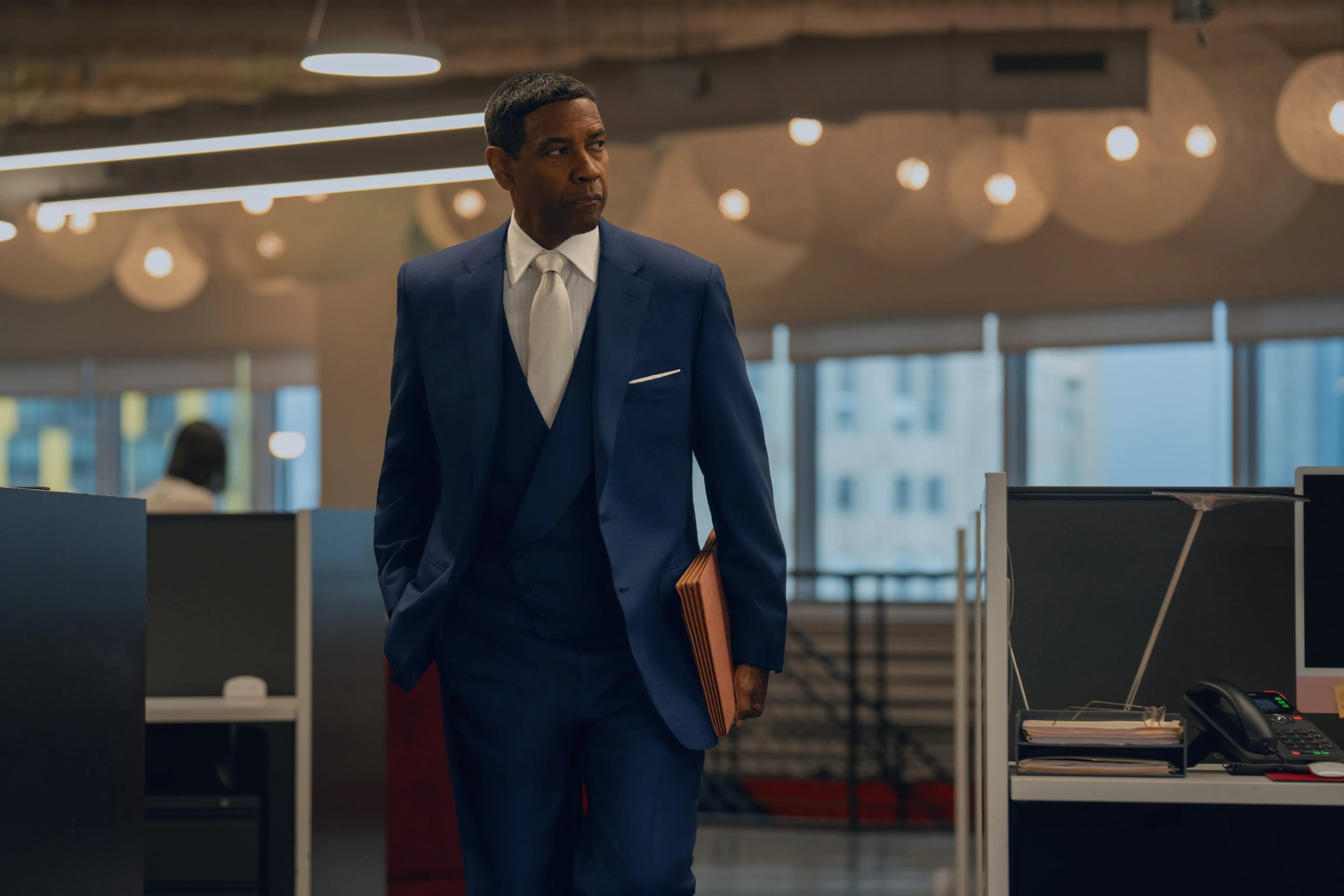 Actor Denzel Washington stands in an office wearing a navy blue suit and carrying a tan briefcase.