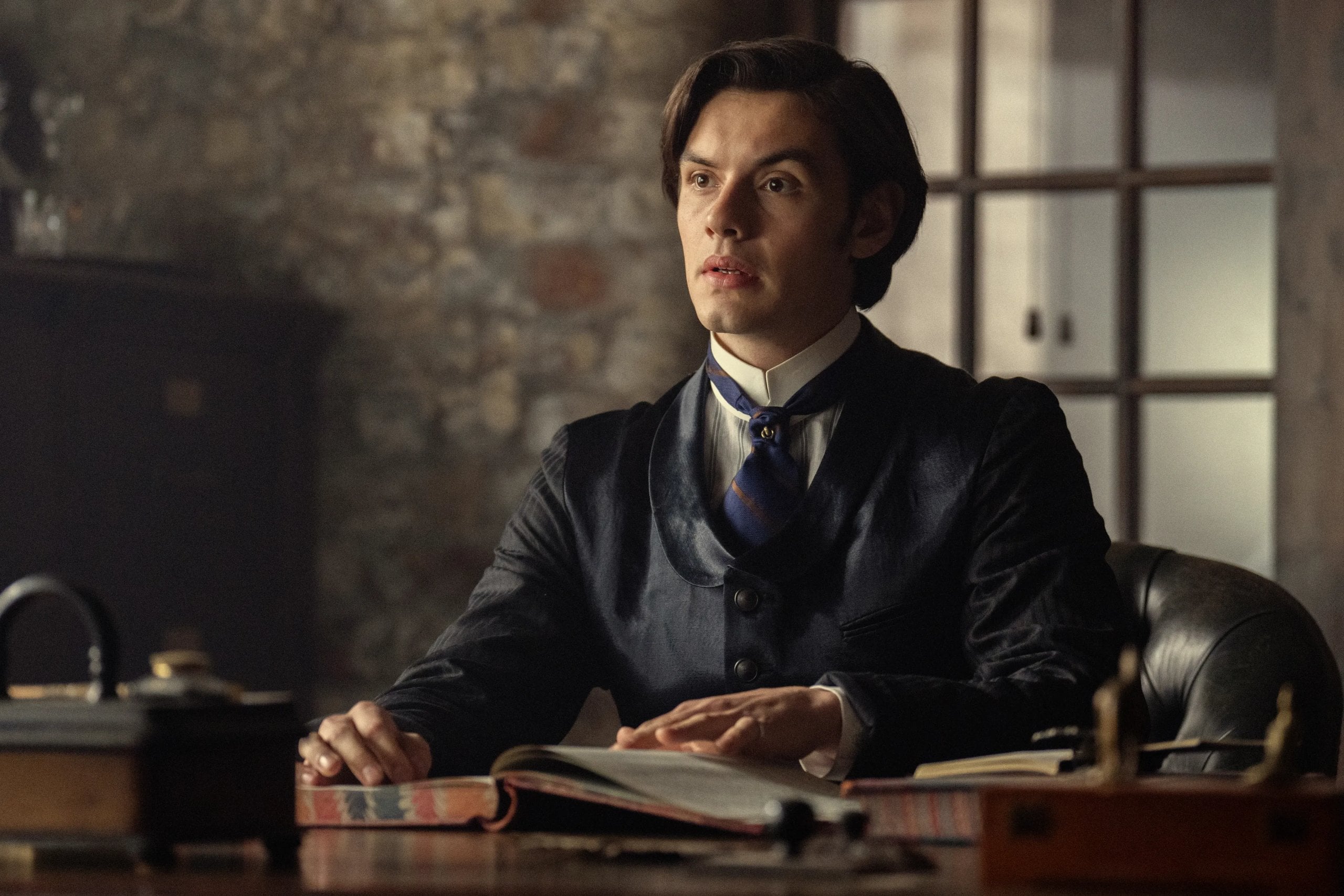 Actor Louis Partidge sits at a desk in a 3-piece, Victoria-era suit in a still from the "House of Guinness."