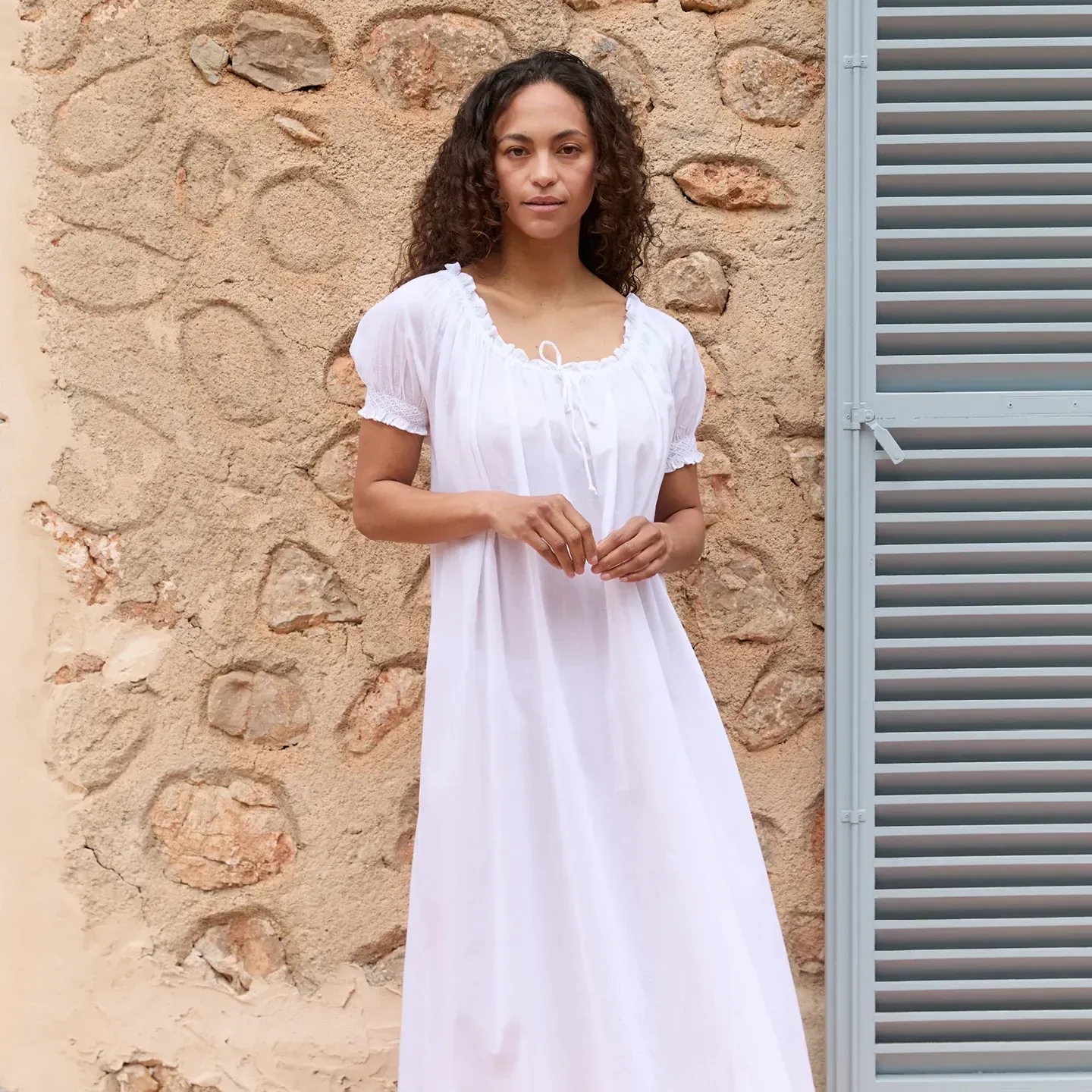 Model standing against a stone wall in a long white nightgown