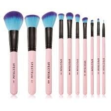 10 Spectrum makeup brushes lined up against a white background, each with a pink handle, black ferrule, and blue and purple bristles