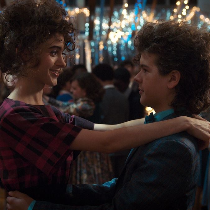 Actors Natalia Dyer and Gaten Matarazzo slow dance at a school dance, dressed in full 80s attire in a still from Stranger Things.