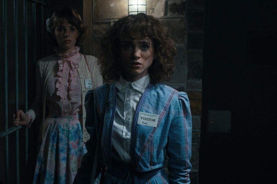 Actors Maya Hawke and Natalia Dyer dressed in 80s-style outfits, walking through a dark hallway in a still from Stranger Things.