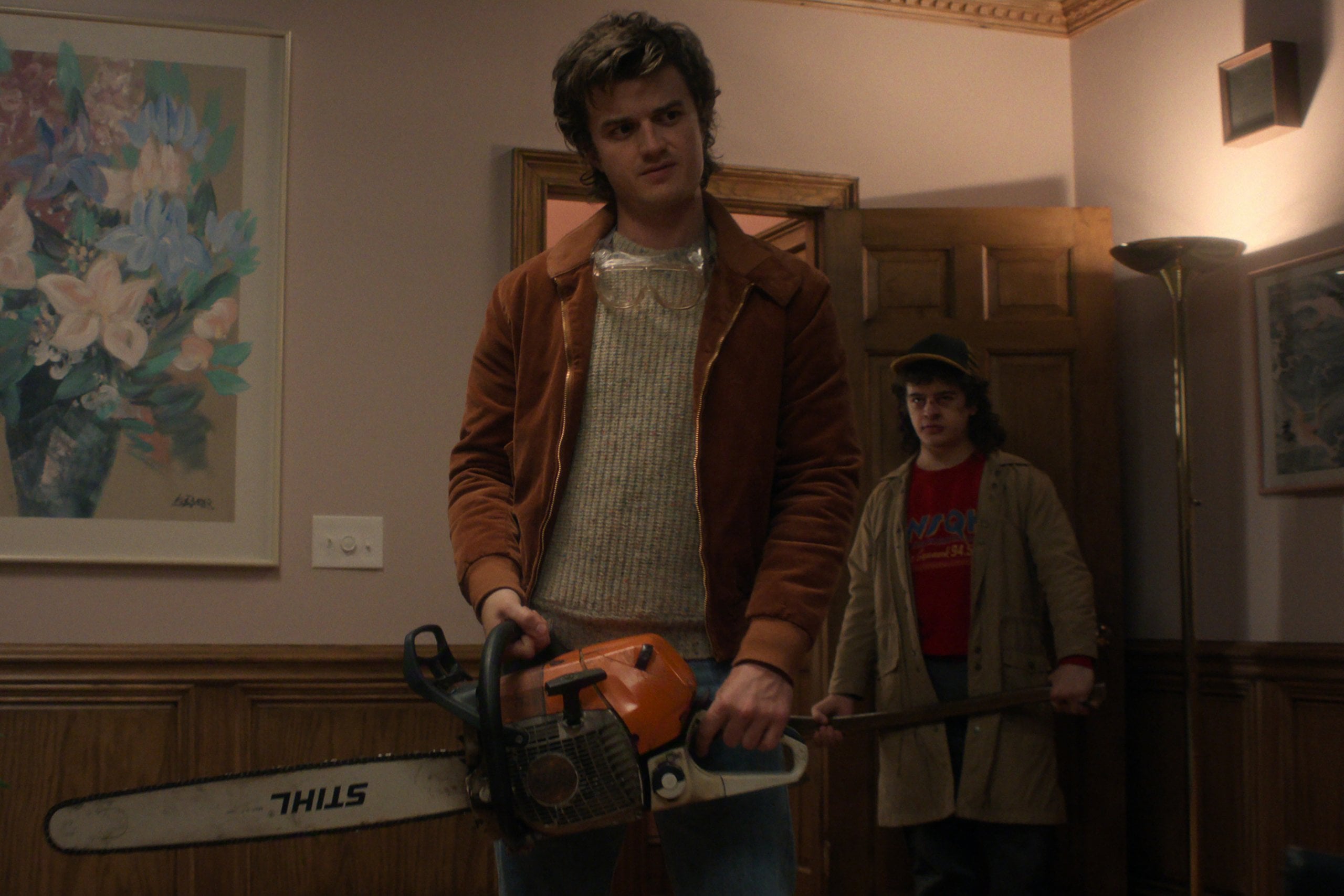 Actor Joe Keery stands in a living room holding a chain saw in "Stranger Things" season five. Photo: Netflix.