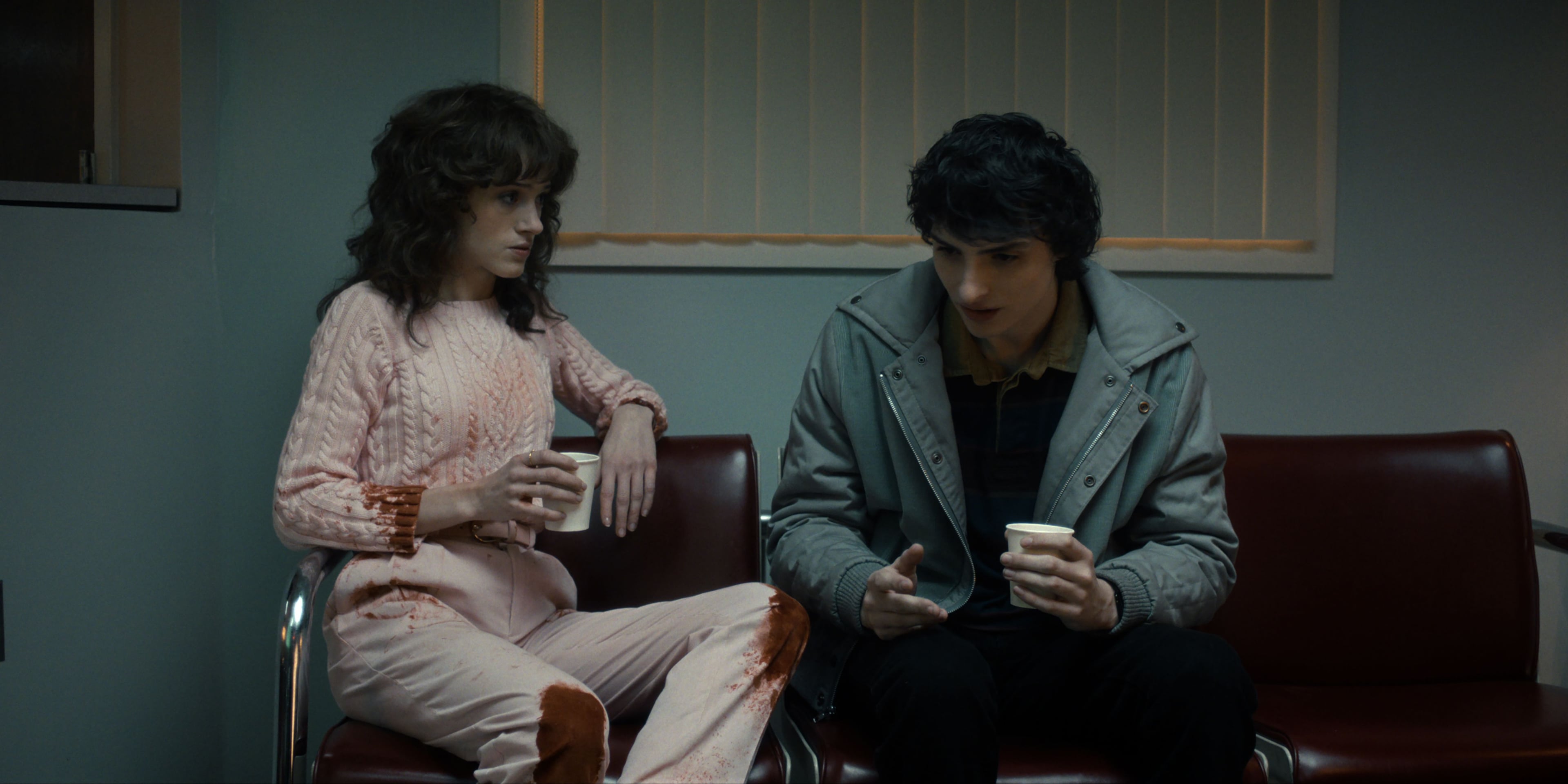 Actors Natalia Dyer and Finn Wolfhard sit next to each other in a hospital lobby in a still from Stranger Things season five.