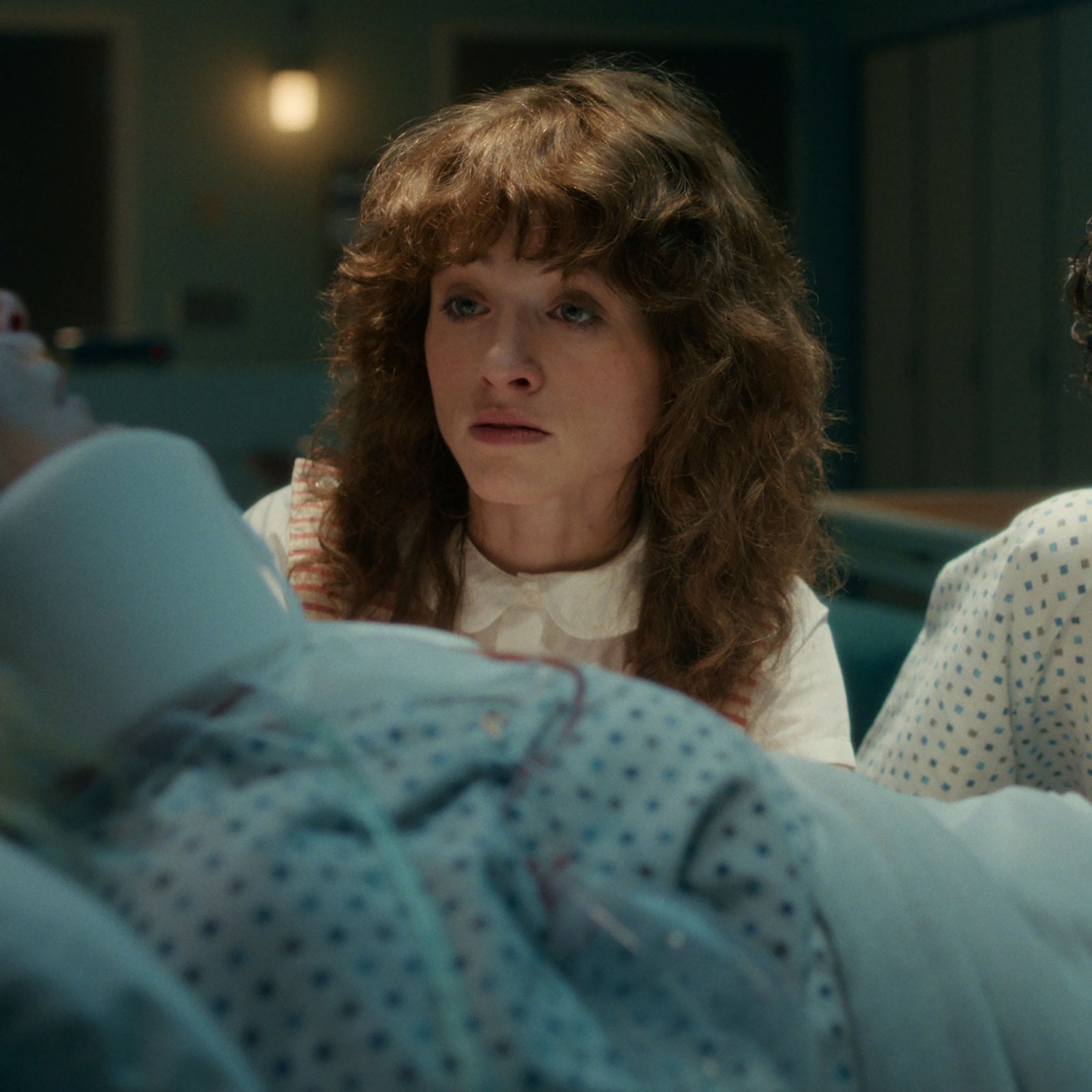 Actors Natalia Dyer and Finn Wolfhard sitting at the side of a hospital bed in a still from season 5 of "Stranger Things."