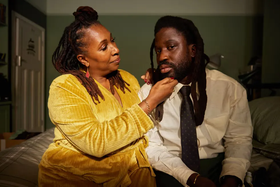 Actor Jo Martin sits on a couch in a yellow short, holding her TV son's chin in a scene from "Dreaming Whilst Black."