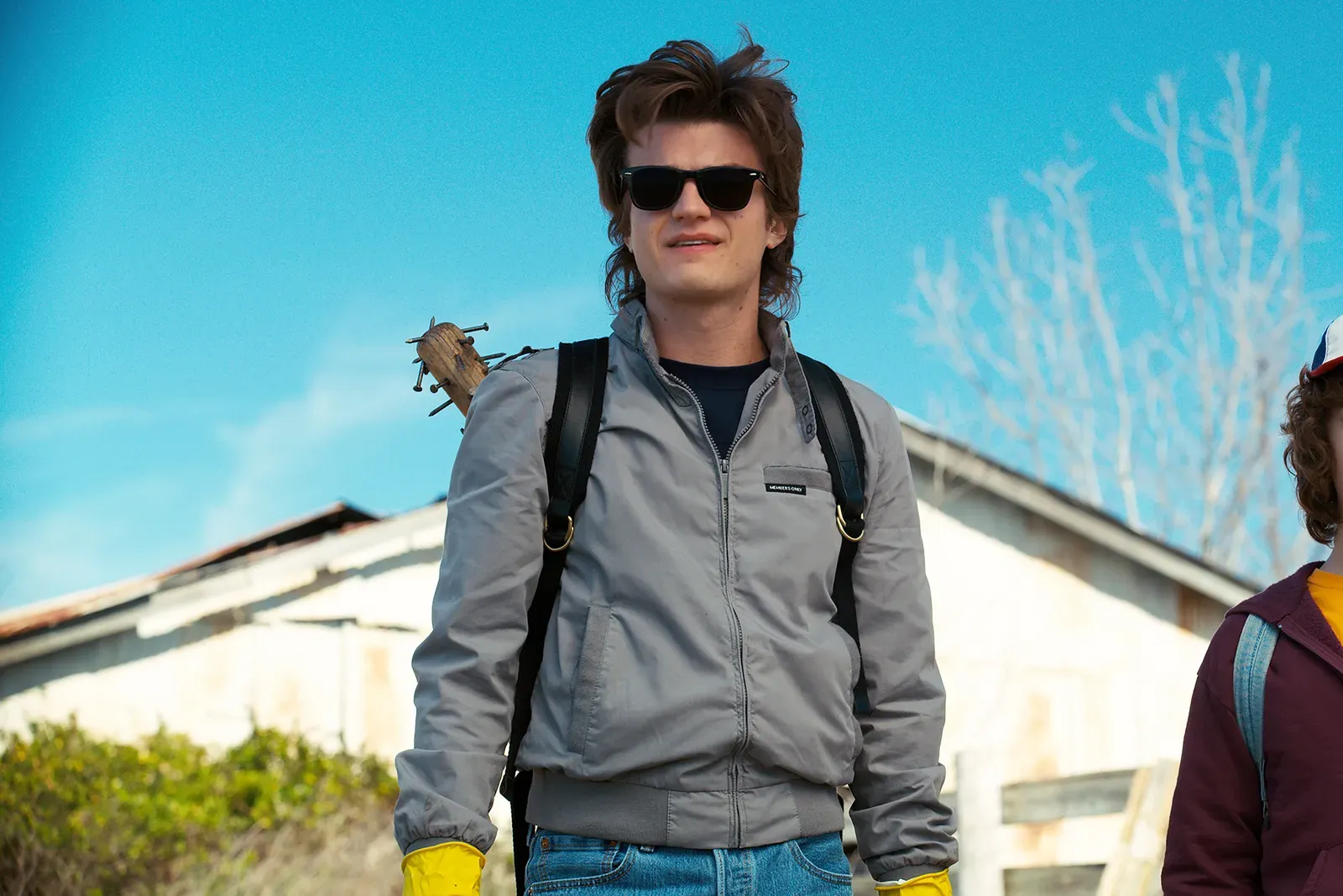 Actor Joe Keery dressed in a gray bomber jacket, wearing a backpack carrying a weapon in a still from Stranger Things.