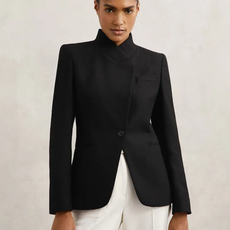 A model wears the Reiss Manon Wool-Blend Wrapover Blazer in Black with white trousers.